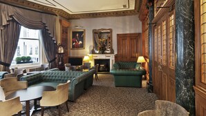 Een klassieke hotel lobby met een groene zithoek, een open haard en een grote spiegel.