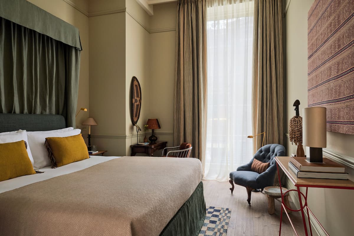 Une chambre d’hôtel avec un grand lit, une chaise, un bureau avec des livres et une fenêtre avec des rideaux.