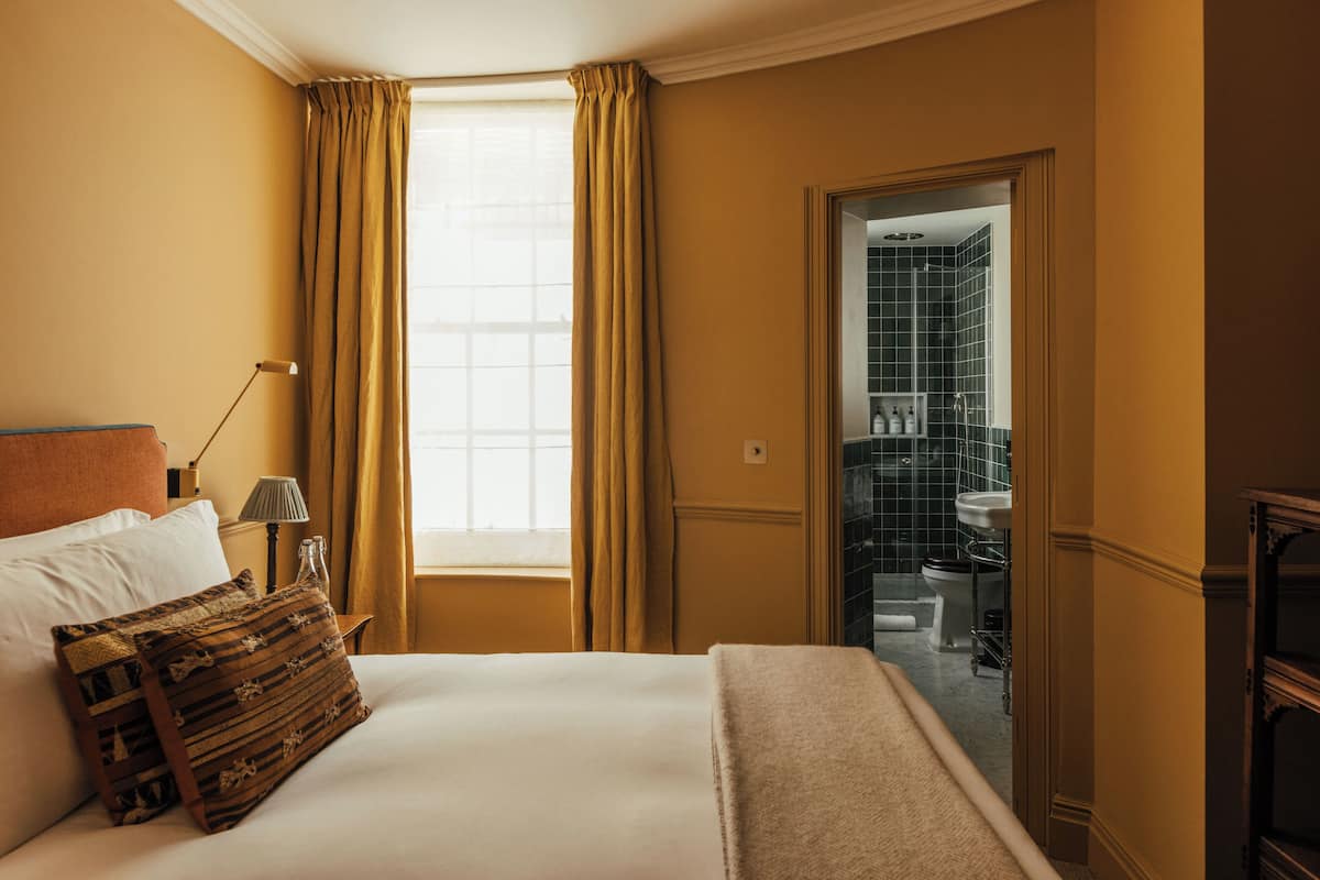 Une chambre d’hôtel avec un lit, une table de chevet, une lampe, une fenêtre avec des rideaux et une vue sur la salle de bain.
