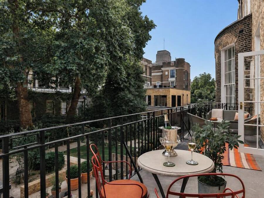Ein Balkon mit Tisch und Stühlen, umgeben von einem Geländer, mit Gebäuden und Bäumen im Hintergrund.