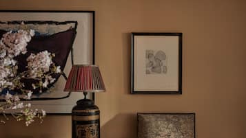 A room with a wooden table, a chair, a lamp, a vase with flowers, and two framed artworks on the wall.