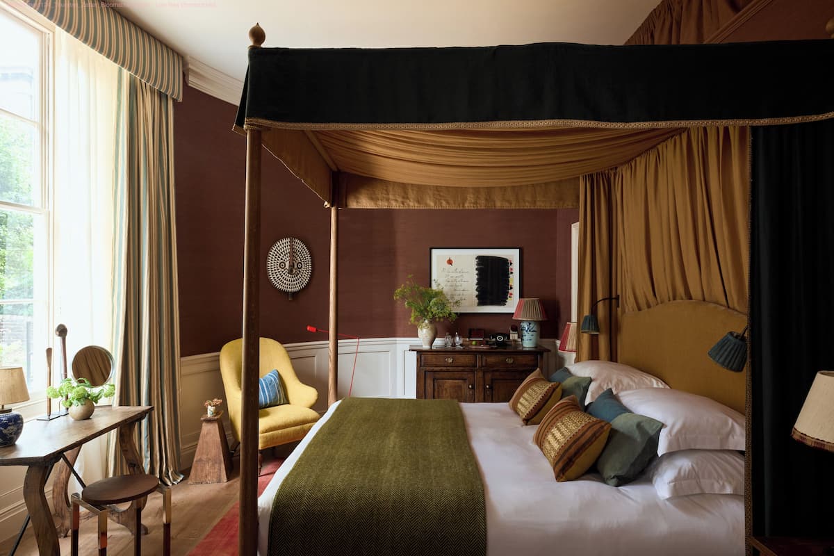 Une chambre avec un lit à baldaquin, un bureau en bois avec une chaise, une petite table avec une lampe et une chaise avec un coussin.