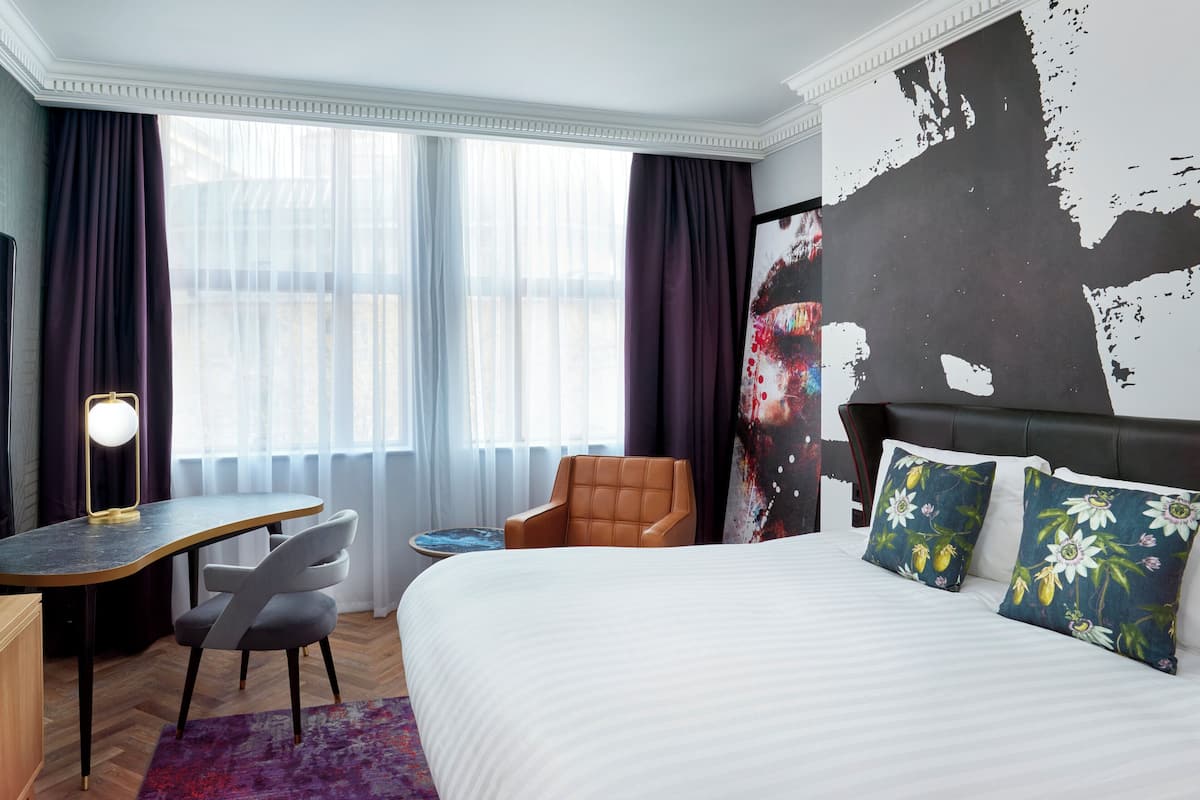 Habitación de hotel con cama, escritorio, silla, televisor y una ventana grande con cortinas.