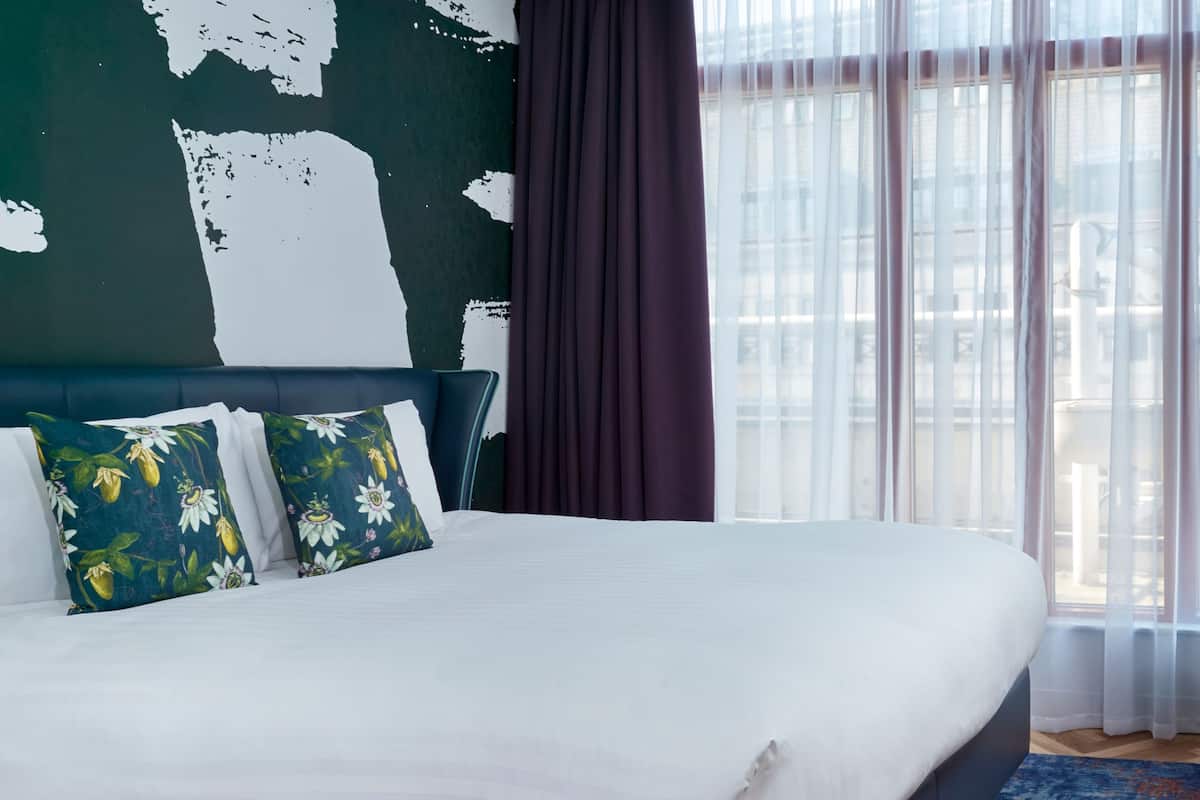 Una cama con ropa de cama blanca, dos almohadas estampadas con flores, un cabecero oscuro y vistas al exterior a través de cortinas de gasa.