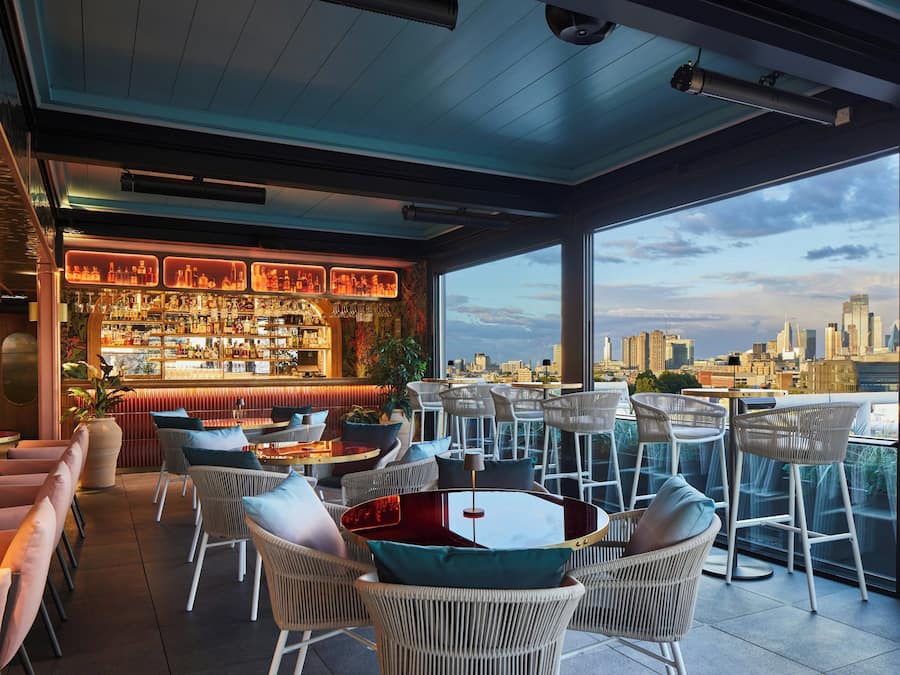 Un bar en la azotea con vista a la ciudad, mobiliario de mimbre y una barra bien surtida.