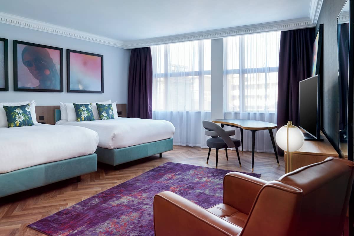 Habitación de hotel con dos camas, un escritorio, una silla, un televisor y ventanas grandes con cortinas.