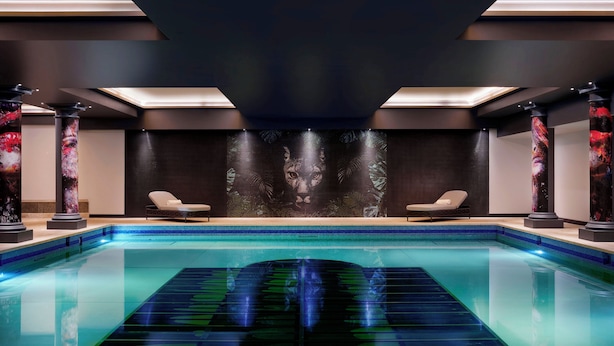 Una piscina interior moderna con agua cristalina, dos sillas de descanso y un gran mural en la pared.