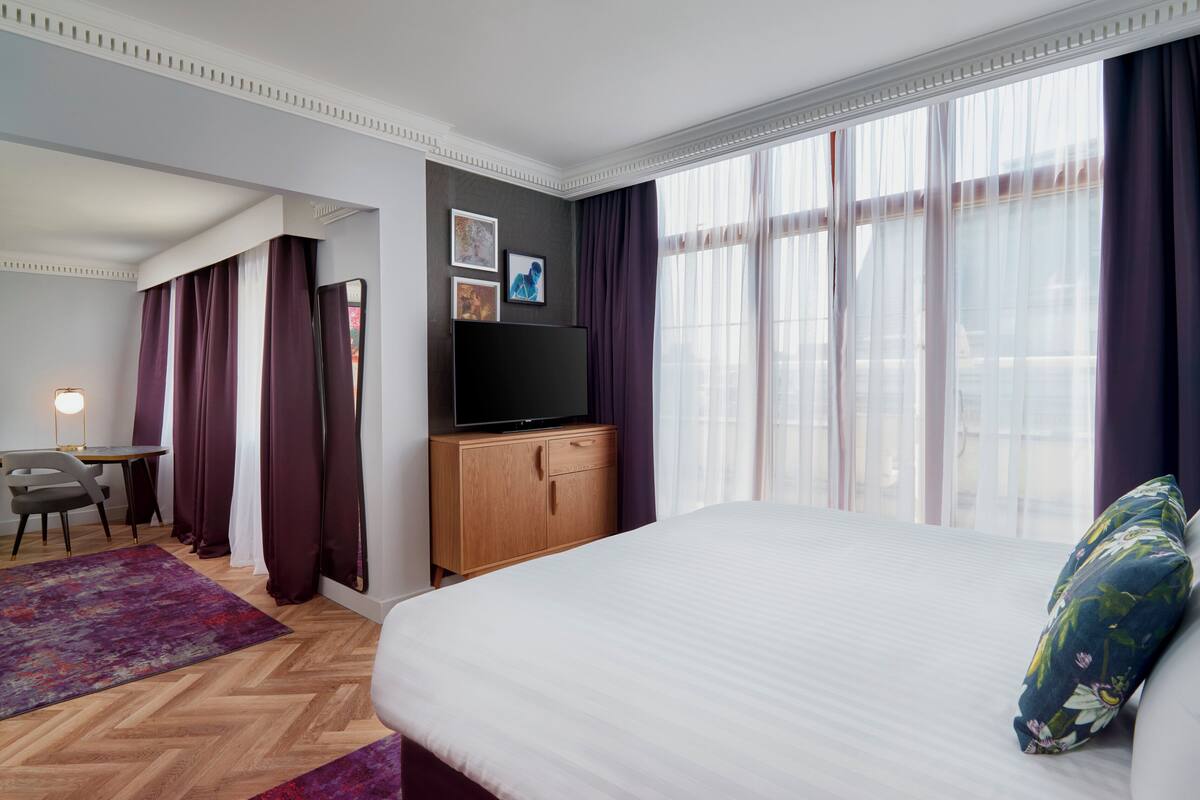 Una habitación de hotel con una cama grande, un escritorio con silla, televisor y ventana con cortinas.