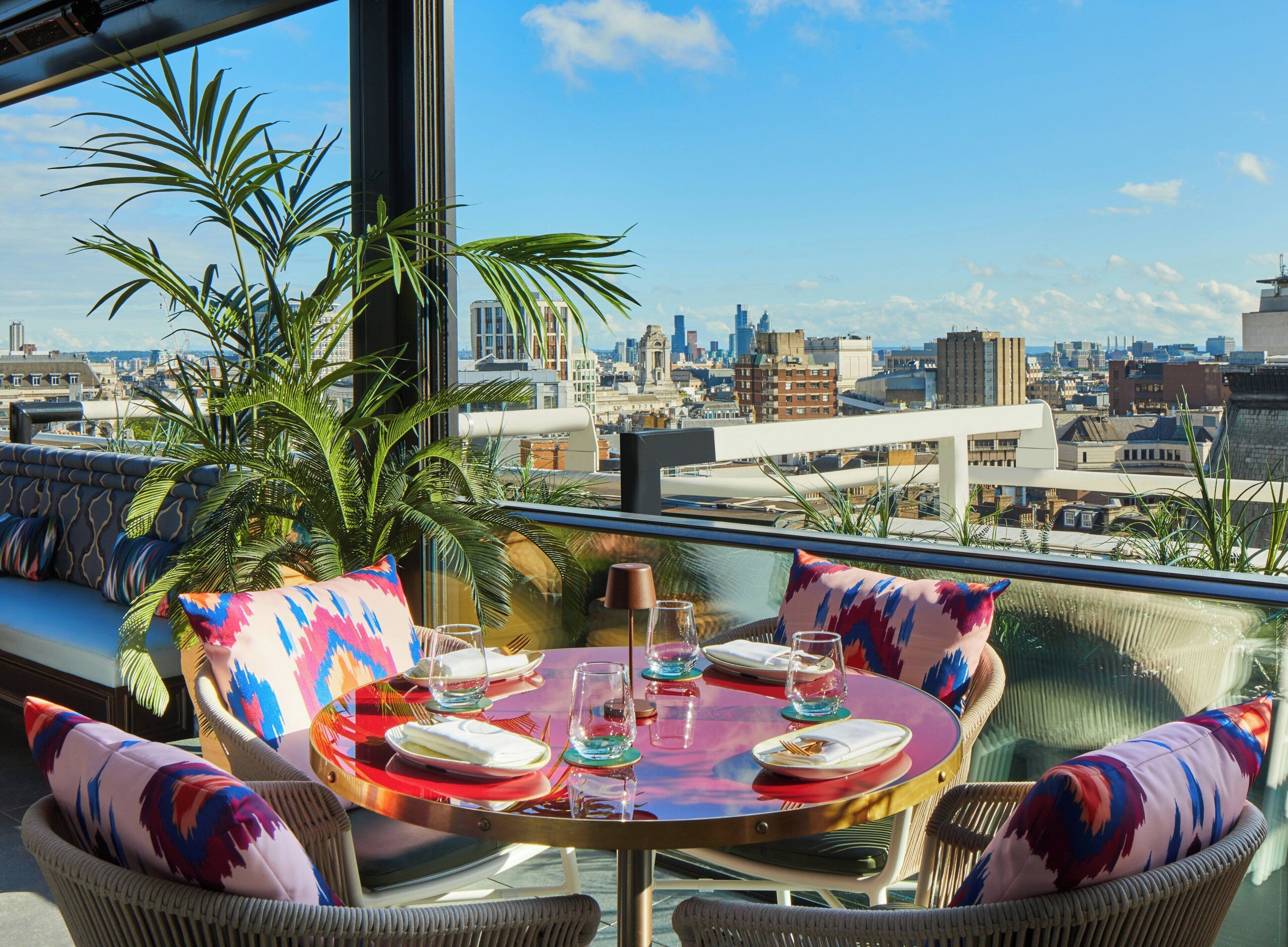 A rooftop dining area with a table set for four, surrounded by wicker chairs with colorful cushions, overlooking a city skyline.