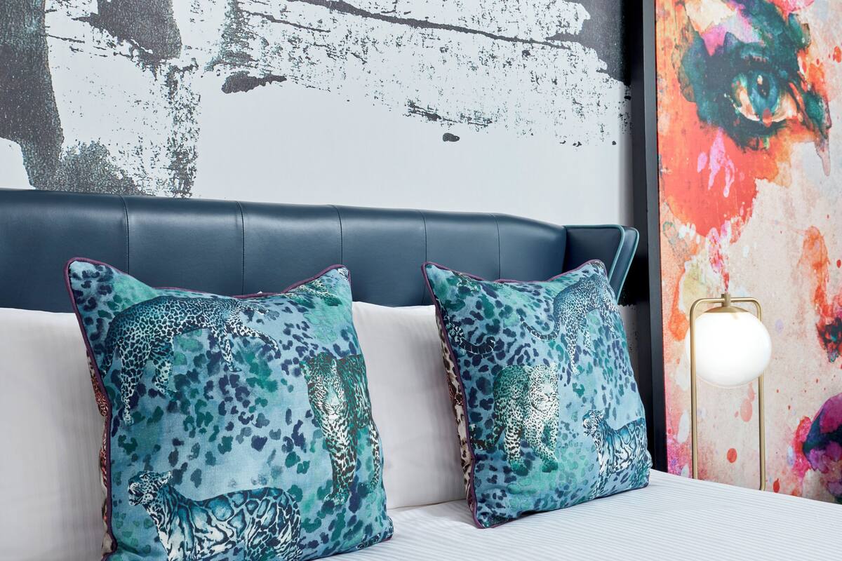 Una cama con dos almohadas de estampado de leopardo, un cabecero oscuro y un cuadro abstracto colorido en la pared.