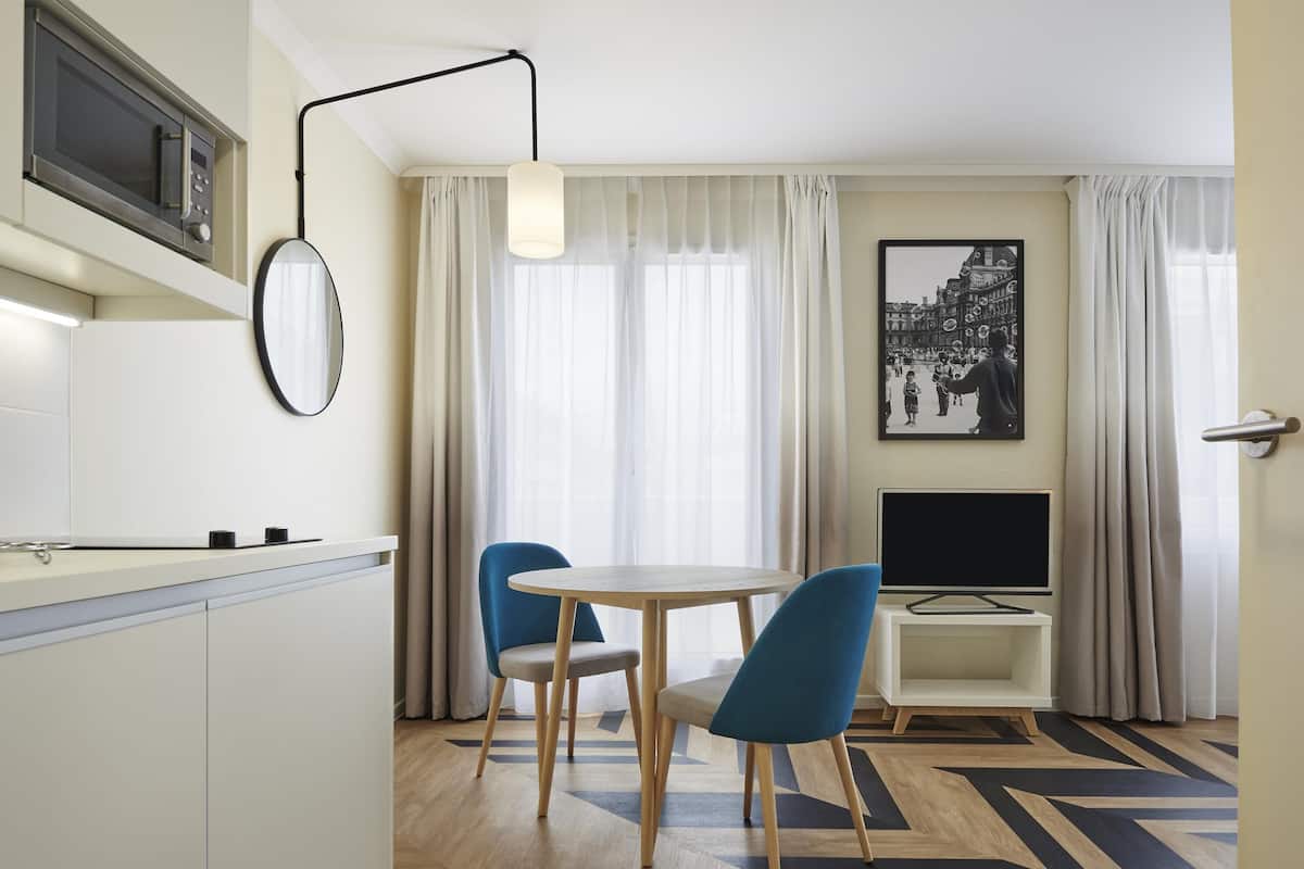 A modern hotel room with a kitchen, a dining area with two chairs, a flat-screen TV, and a large window with sheer curtains.