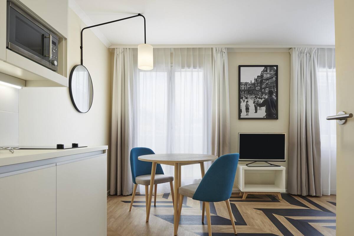 Une chambre d’hôtel moderne dotée d’une cuisine, d’un coin repas avec deux chaises, d’un téléviseur à écran plat et d’une grande fenêtre agrémentée de rideaux légers.