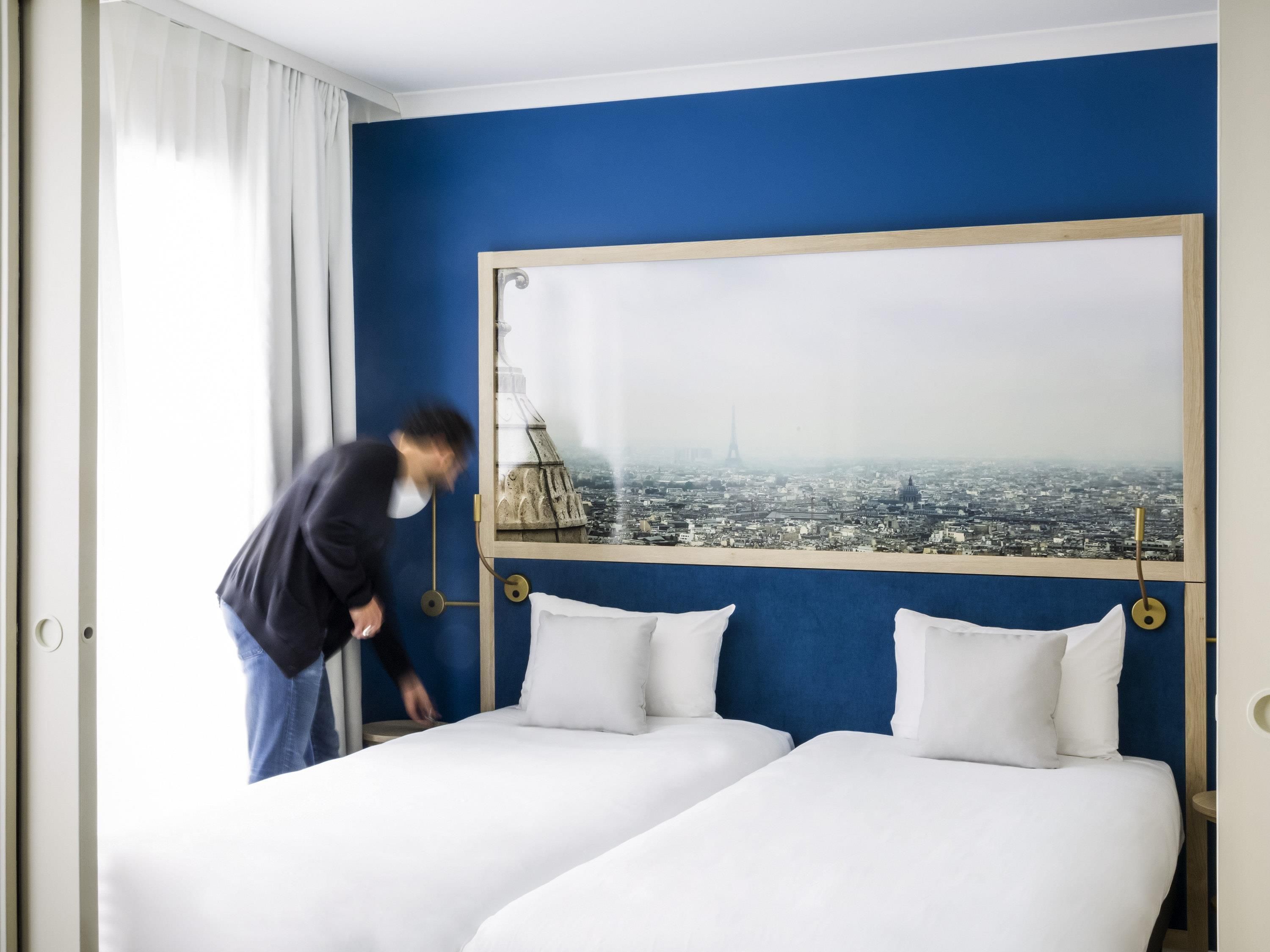A hotel room with two beds, a large window with a cityscape view, and a curtain on the left.