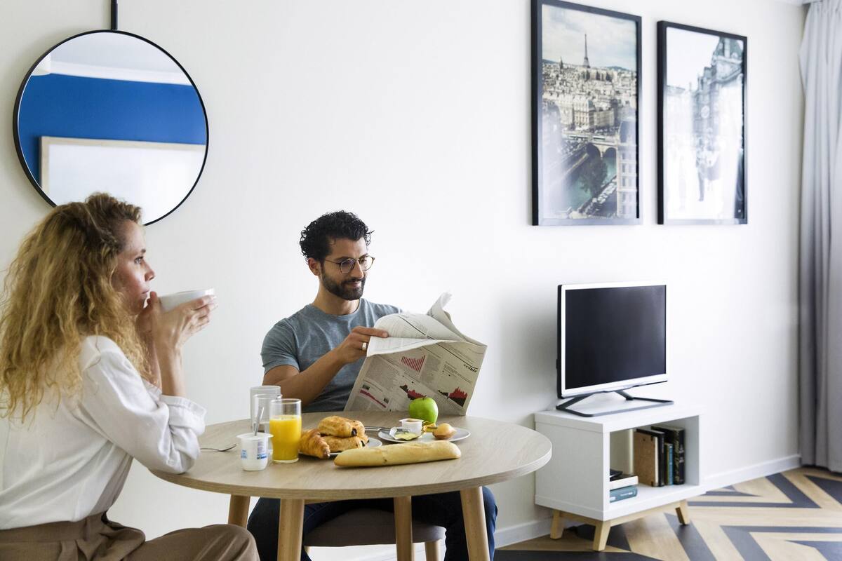 Une chambre d’hôtel moderne avec une table de salle à manger ronde, des articles pour le petit-déjeuner, un téléviseur à écran plat et une œuvre d’art représentant un paysage urbain accrochée au mur.