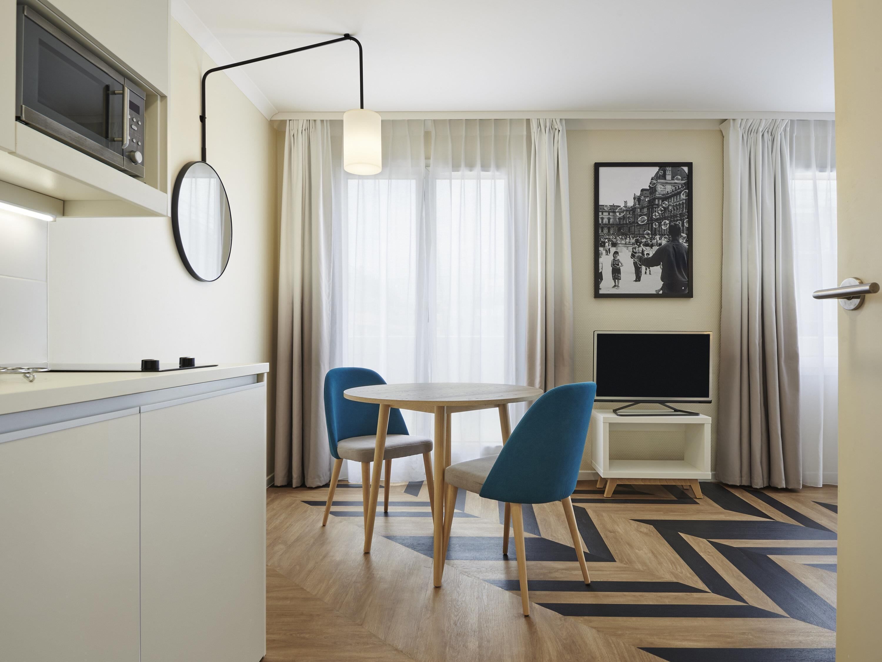 Une chambre d’hôtel moderne dotée d’une cuisine, d’un coin repas avec deux chaises, d’un téléviseur à écran plat et d’une grande fenêtre agrémentée de rideaux légers.