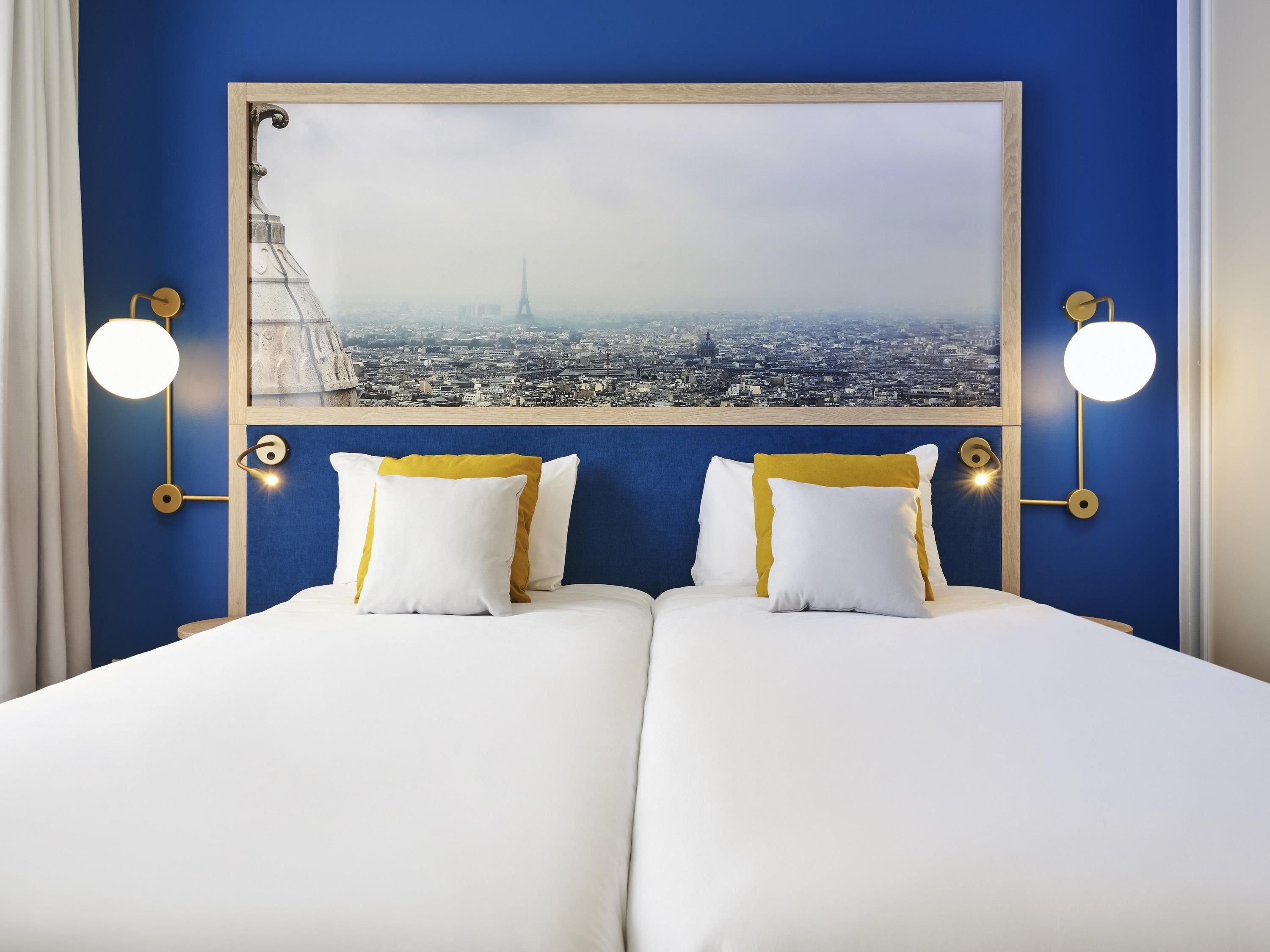 Un lit double avec une literie blanche et jaune, une grande peinture représentant un paysage urbain encadrée au-dessus, et des lampes murales de chaque côté.