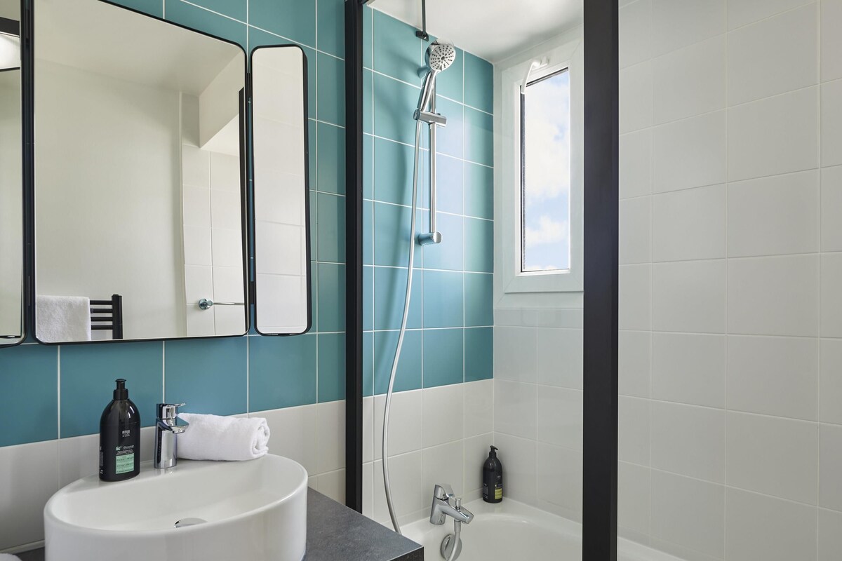 Une salle de bain avec un lavabo blanc, un miroir, une douche et une fenêtre.