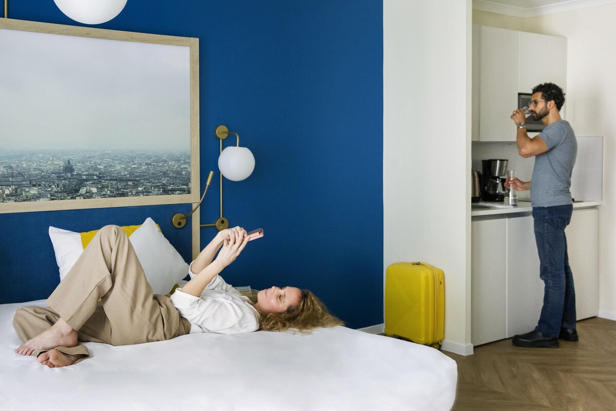 Une chambre avec un lit, une vue sur la ville, un coin cuisine et une valise jaune.