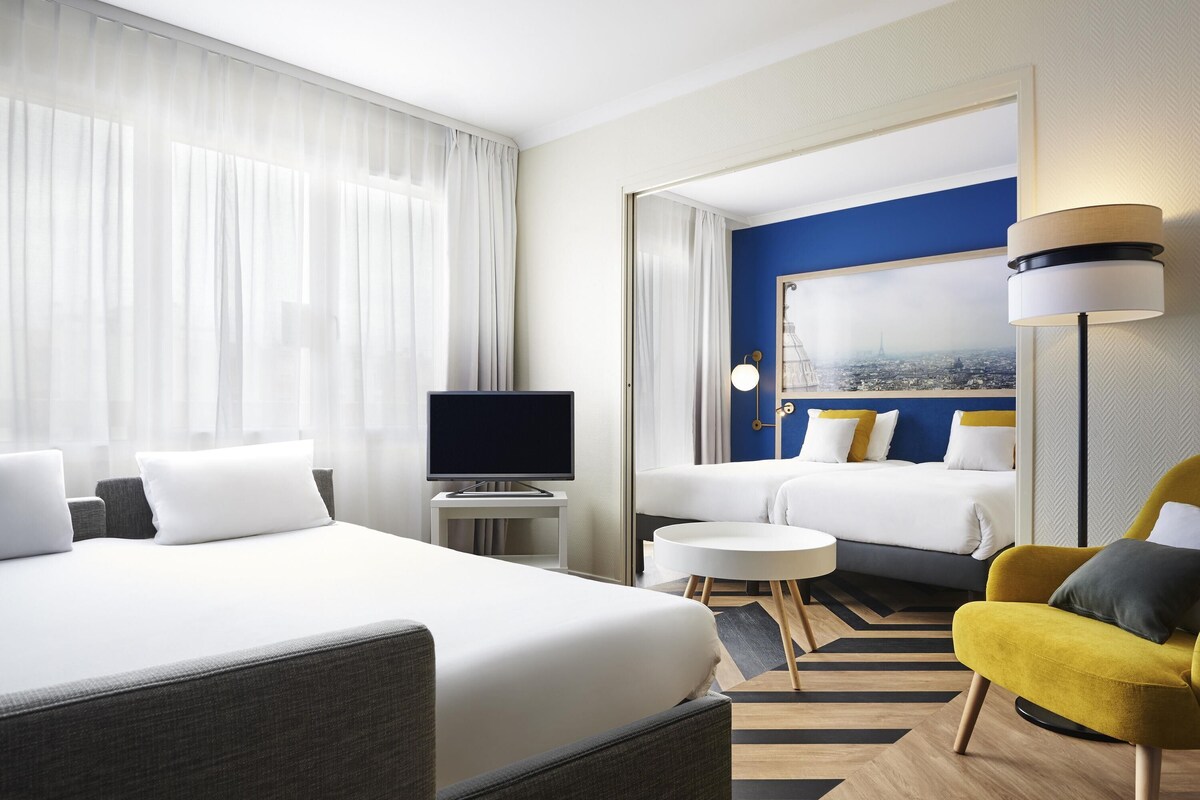 Una habitación de hotel con una cama grande, un escritorio con televisor de pantalla plana, una mesita redonda pequeña, un sillón amarillo y un cuadro de paisaje urbano en la pared.