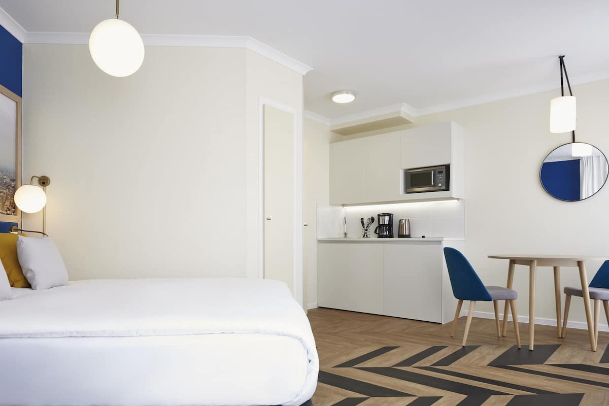 A hotel room with a bed, a small kitchenette, a dining area with chairs, and a mirror on the wall.