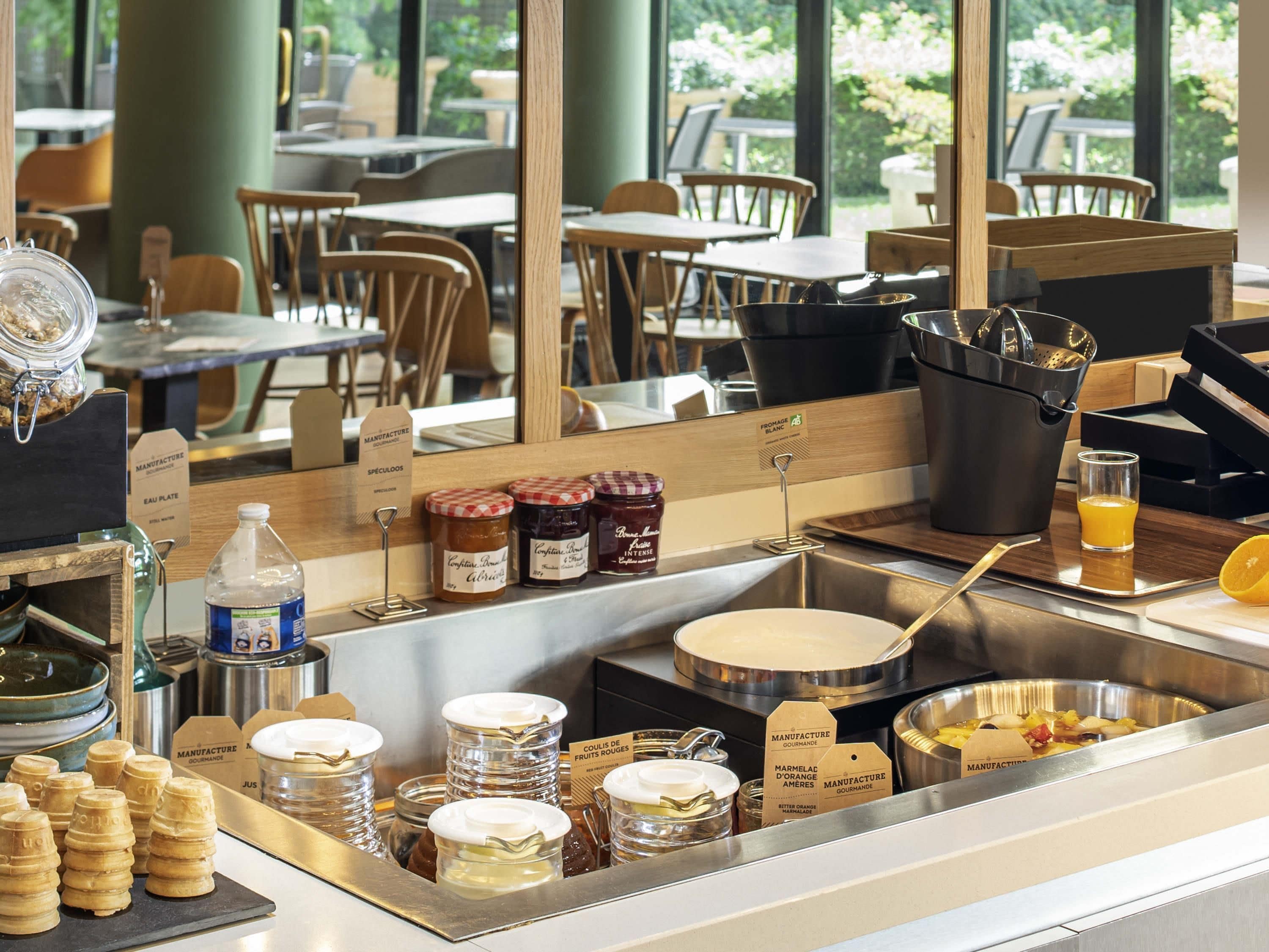 A breakfast buffet area with various food items, a juice dispenser, and a view of the outdoor seating area.