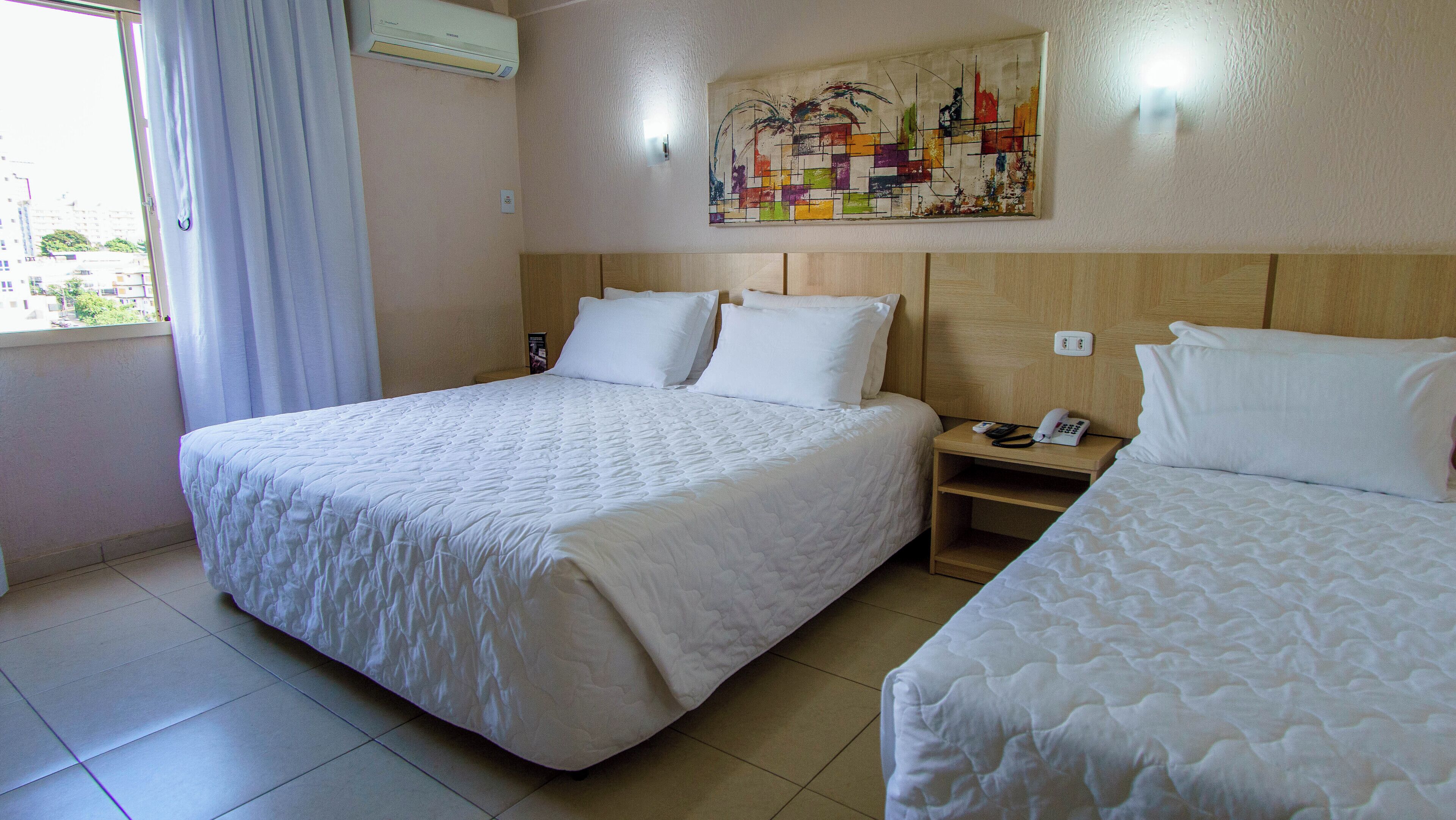 Una habitación de hotel con una cama grande, una cama más pequeña, una mesita de noche con un teléfono y un cuadro en la pared.