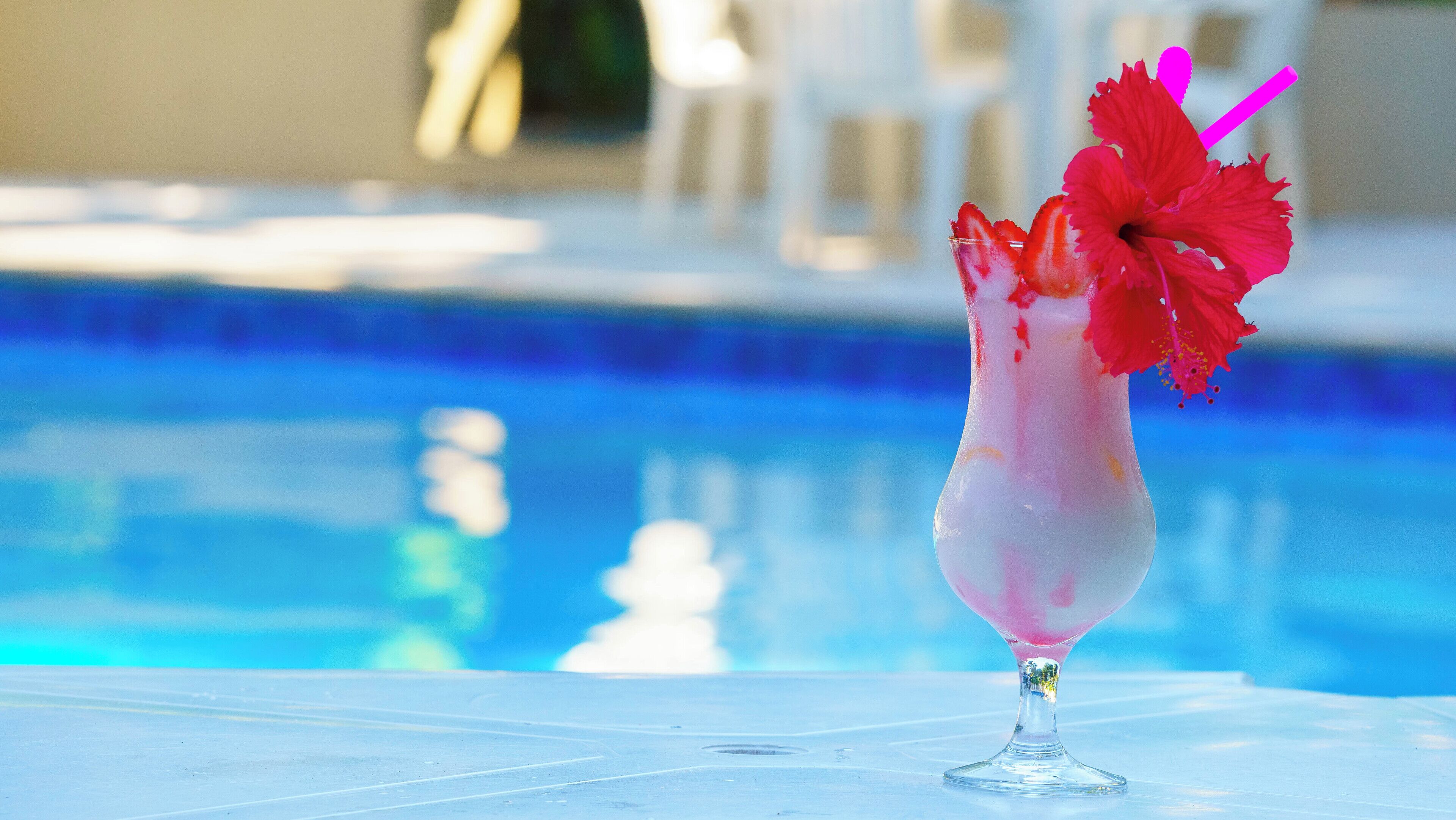 Een cocktail in een glas met een roze tint en een rode bloem als garnering, geplaatst op een tafel bij een zwembad.