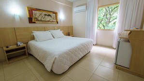 A hotel room with a large bed, a bedside table with a phone, a small cabinet, a window with curtains, and a wall-mounted air conditioner.