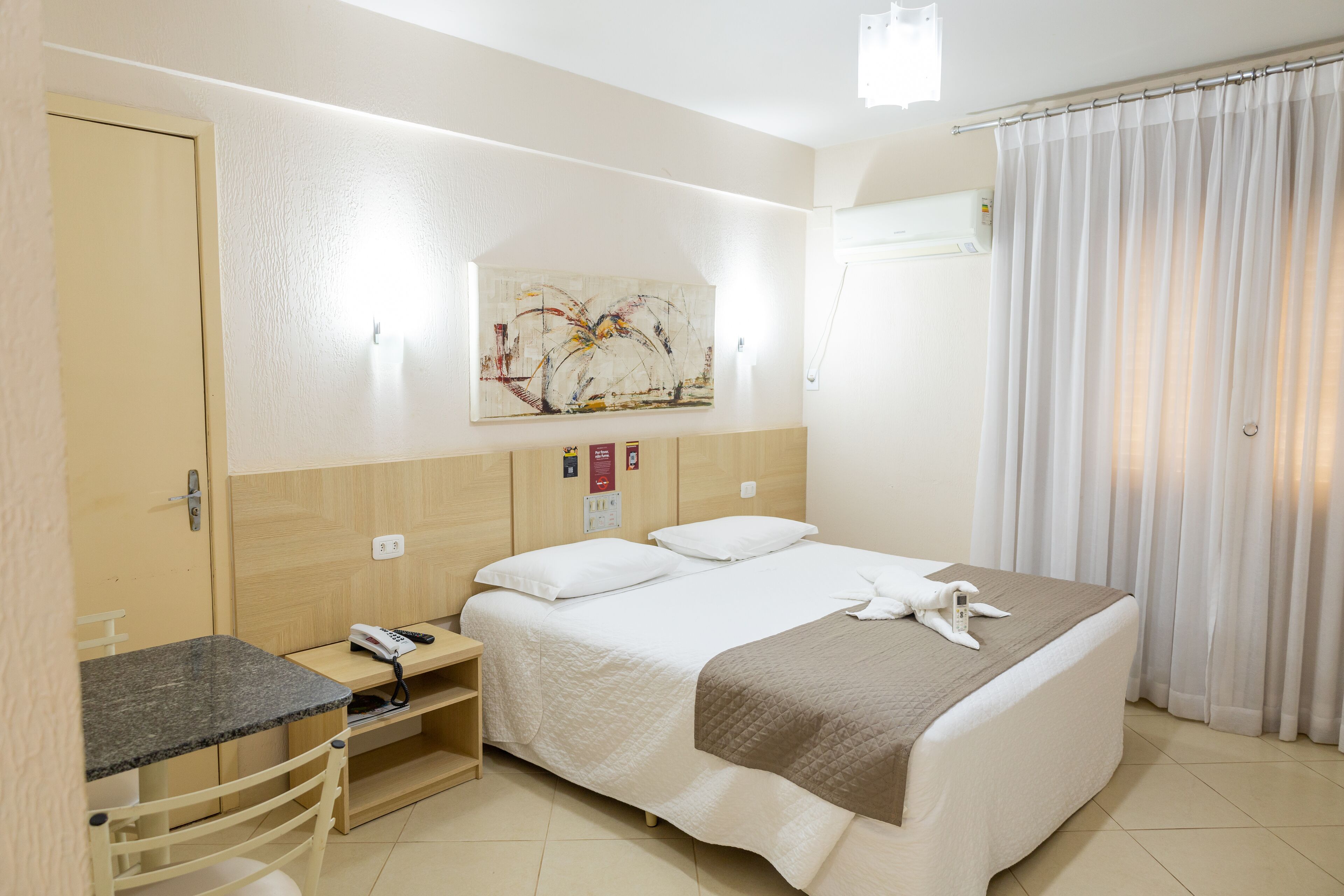 Quarto de hotel com cama, escrivaninha, cadeira, telefone e um quadro na parede.