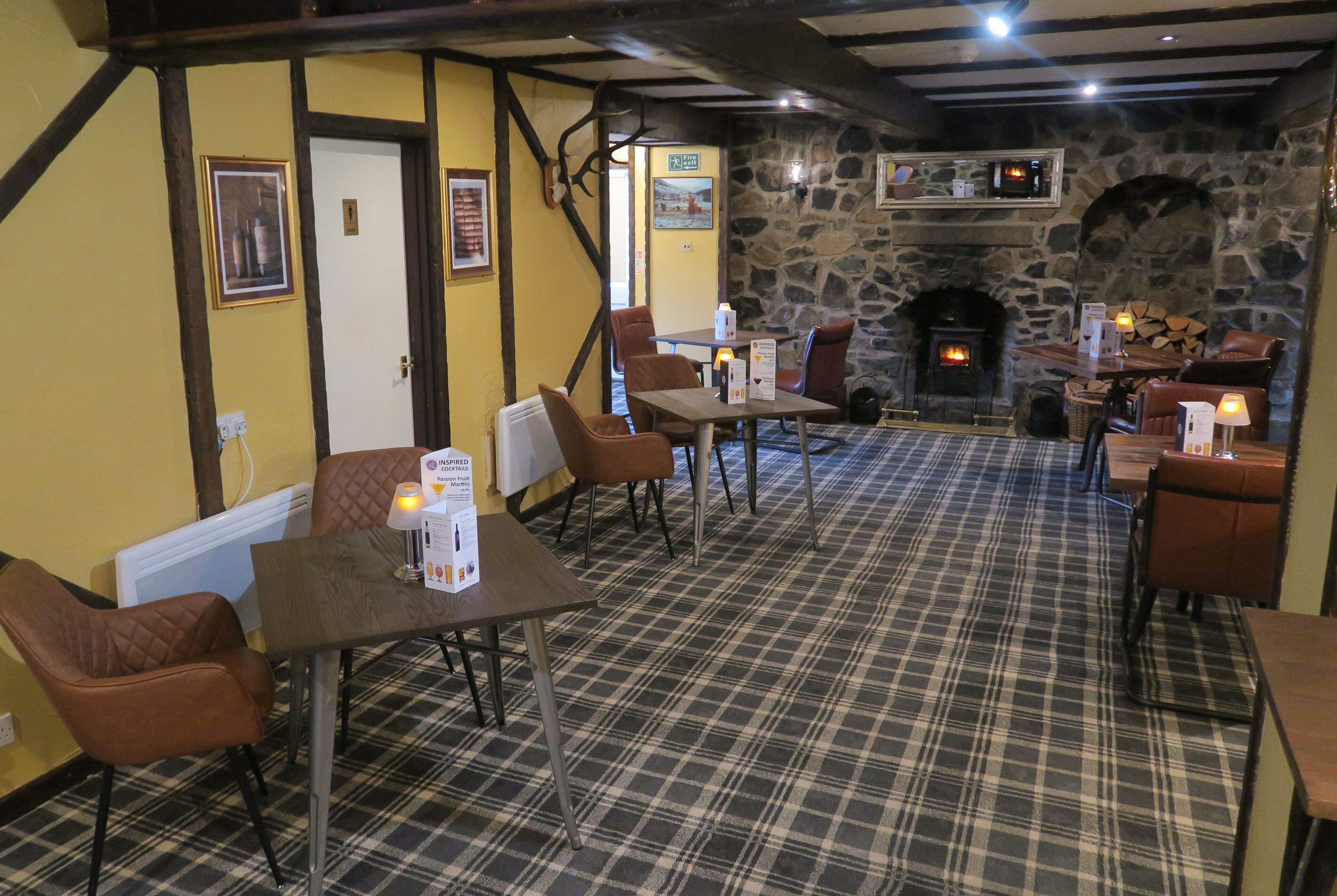 A cozy restaurant with a stone fireplace, plaid carpet, and wooden beams. Tables and chairs are arranged for dining.