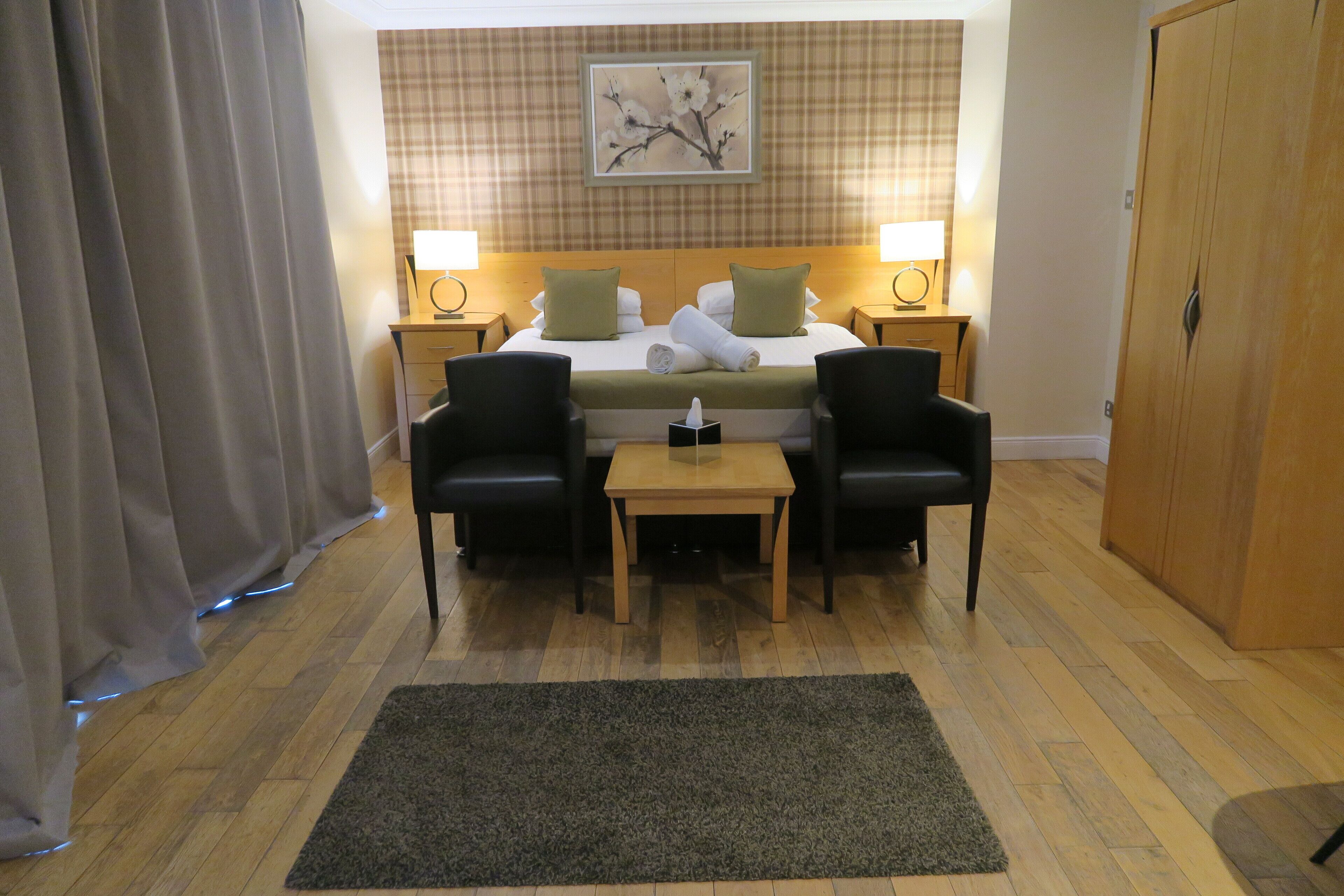 A hotel room with a bed, two bedside tables with lamps, a small table with a tissue box, two armchairs, a dark rug, wooden flooring, and a wardrobe.