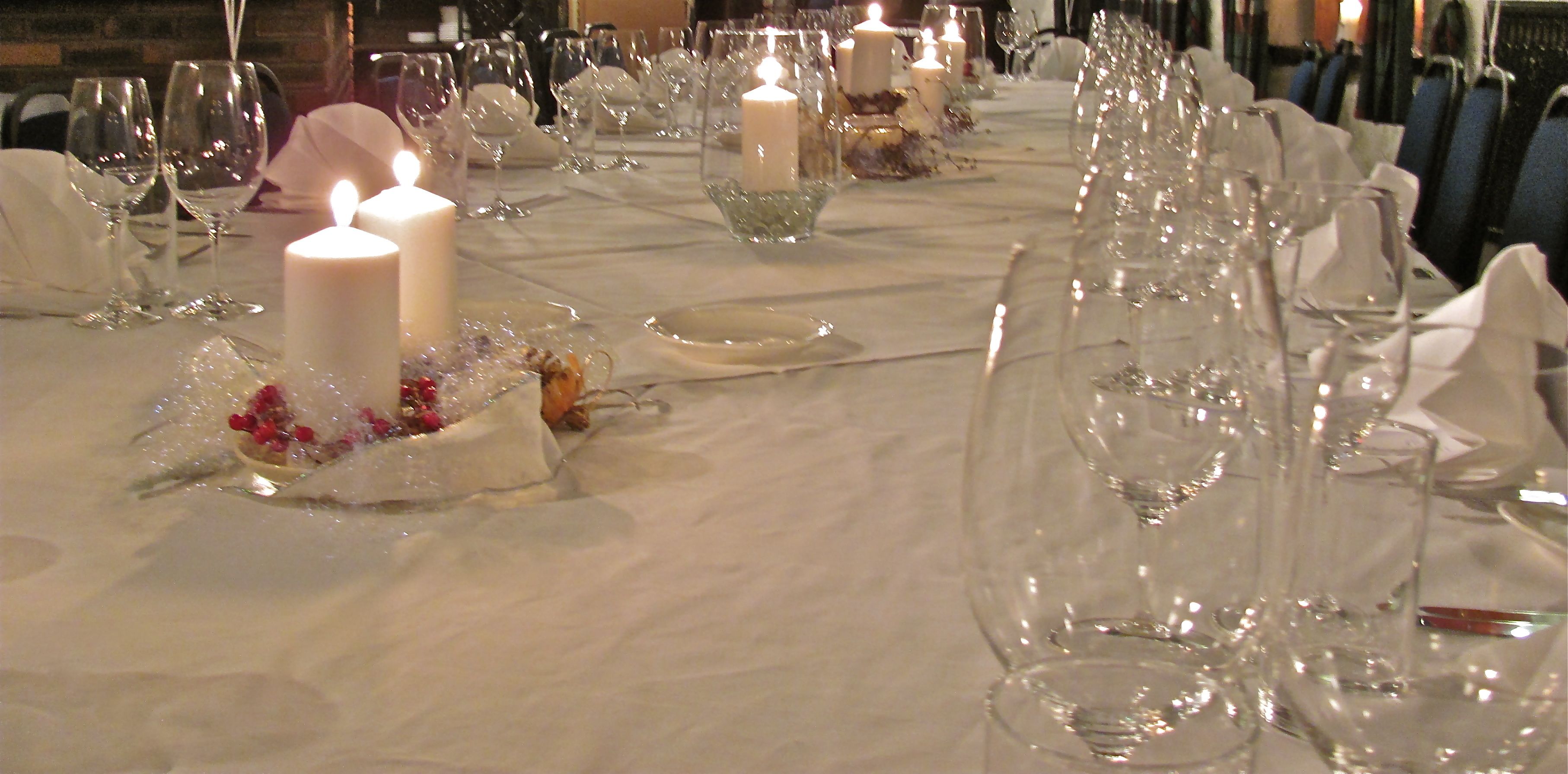 Una larga mesa de comedor dispuesta para un evento formal, con velas, cristalería transparente y manteles blancos.