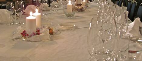 Una larga mesa de comedor dispuesta para un evento formal, con velas, cristalería transparente y manteles blancos.