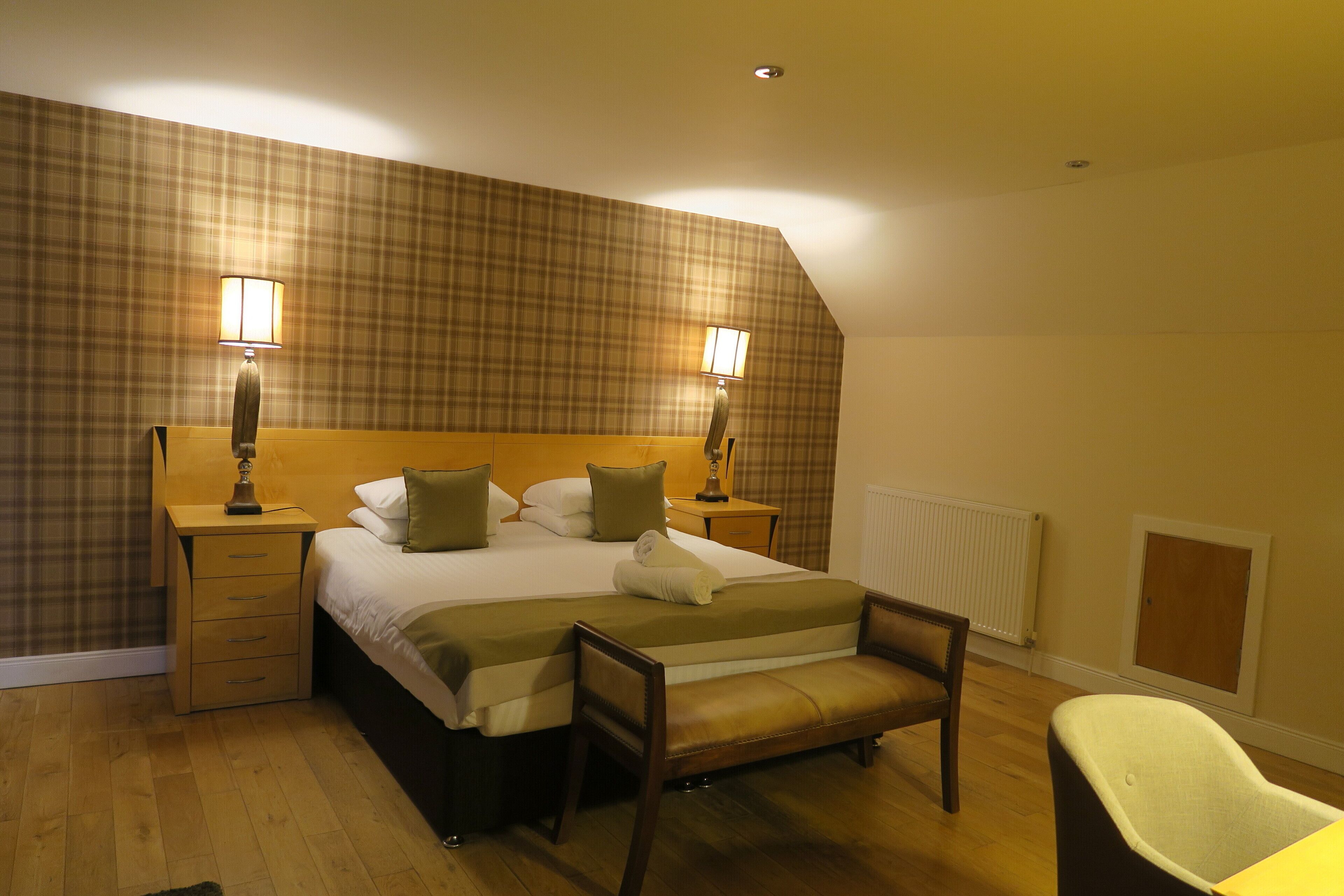 A hotel room with a large bed, two bedside lamps, a wooden nightstand, a bench, and a chair.