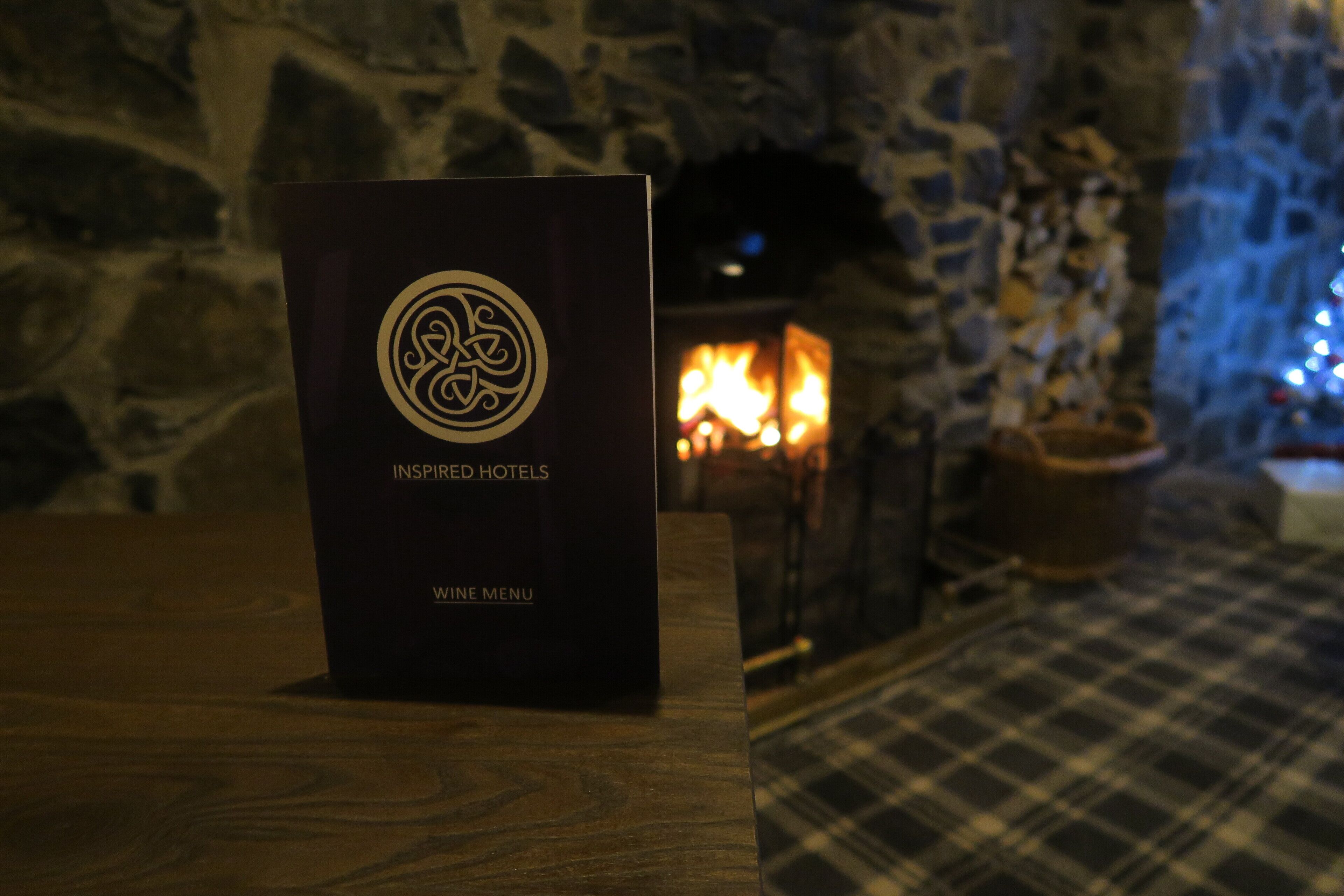 A black sign with a logo and text reading "INSPIRED HOTELS" and "WINE MENU" on a wooden table in a room with a stone wall and a fireplace in the background.