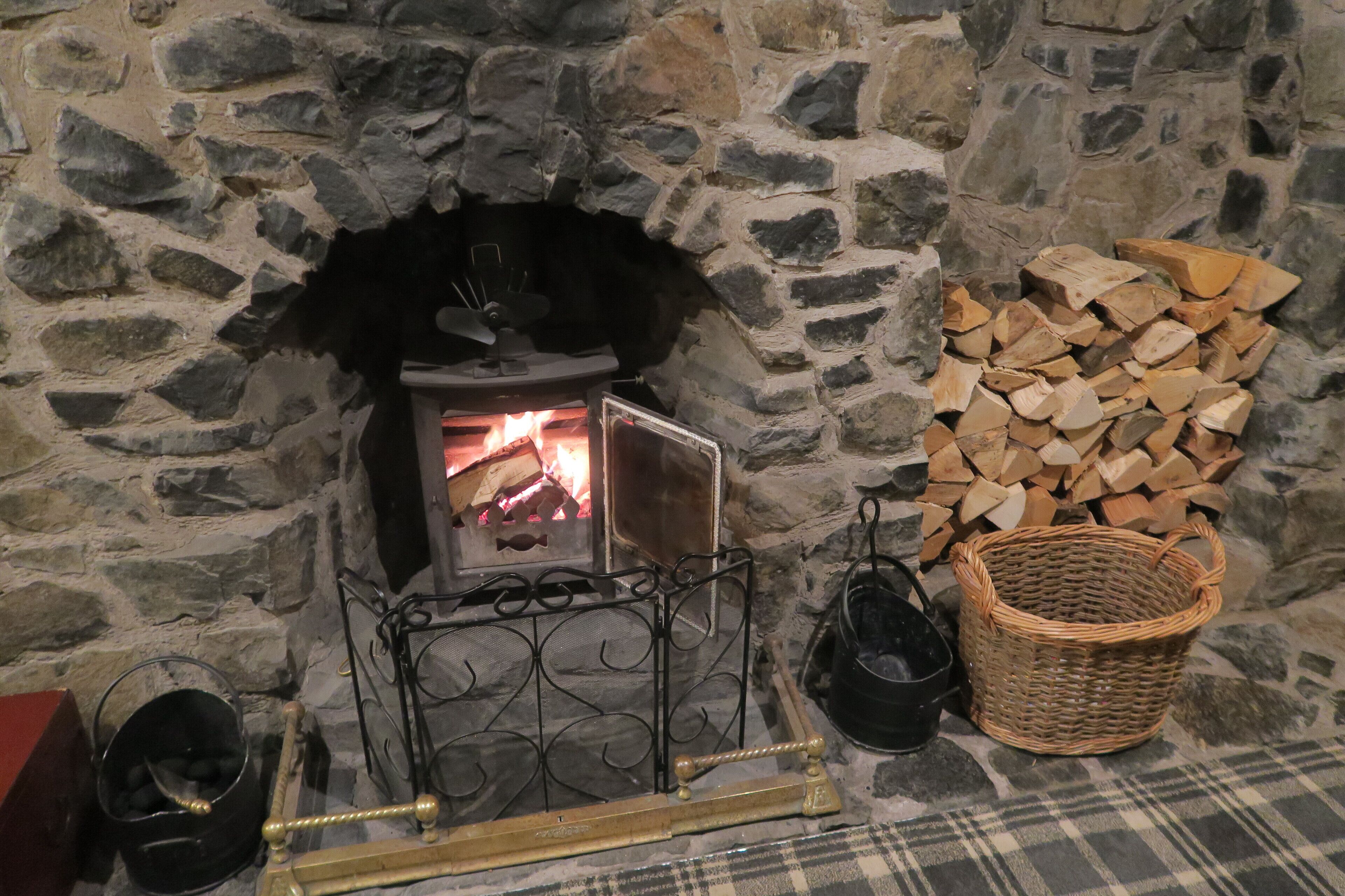 Une cheminée en pierre avec un feu de bois, un foyer en métal, un panier et une pile de bois de chauffage.