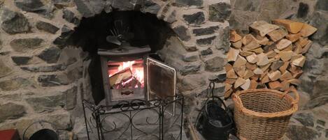 Une cheminée en pierre avec un feu de bois, un foyer en métal, un panier et une pile de bois de chauffage.