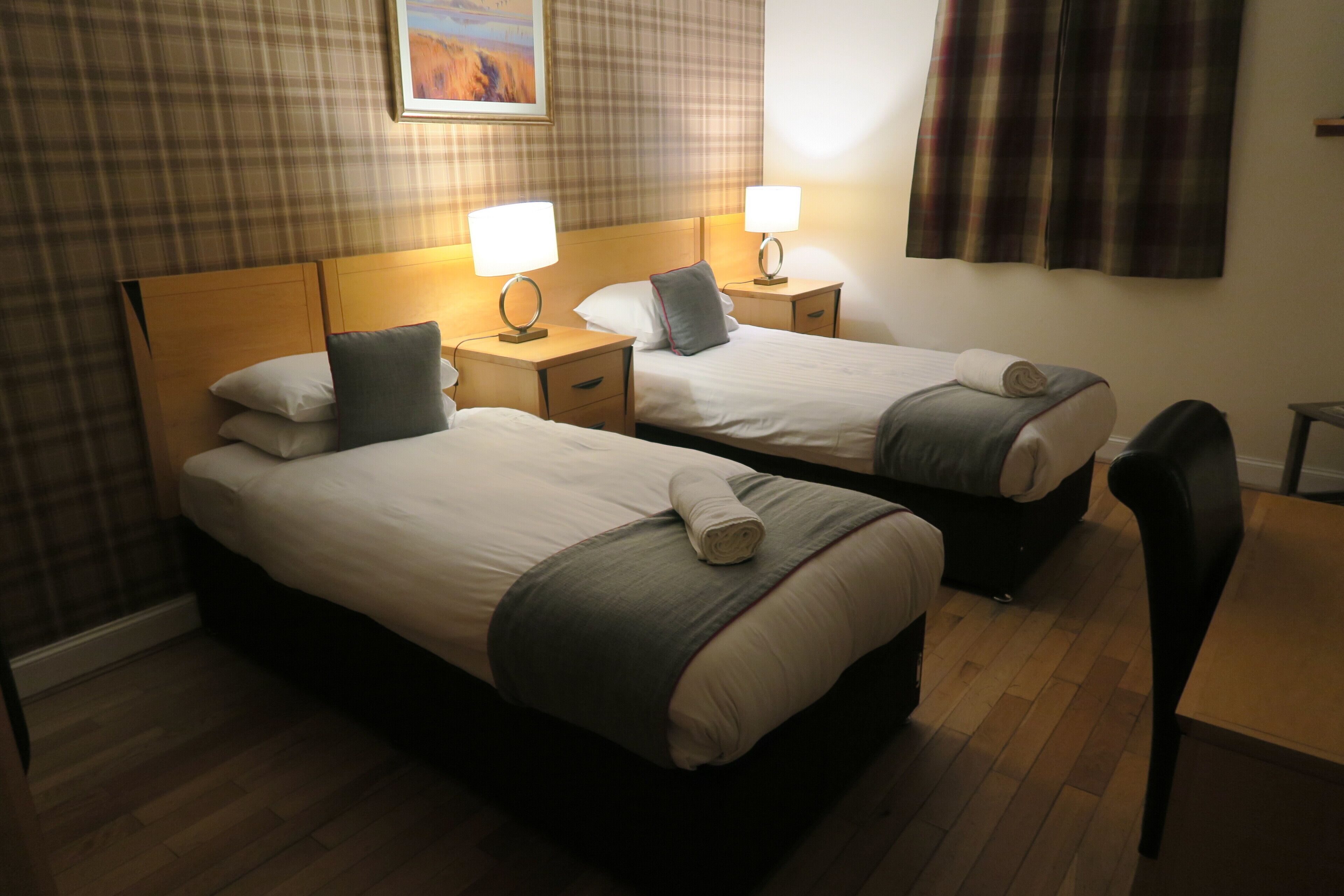 A hotel room with two single beds, each with a pillow and a rolled towel. There are bedside tables with lamps, a wooden floor, and a chair.