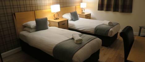 A hotel room with two single beds, each with a pillow and a rolled towel. There are bedside tables with lamps, a wooden floor, and a chair.