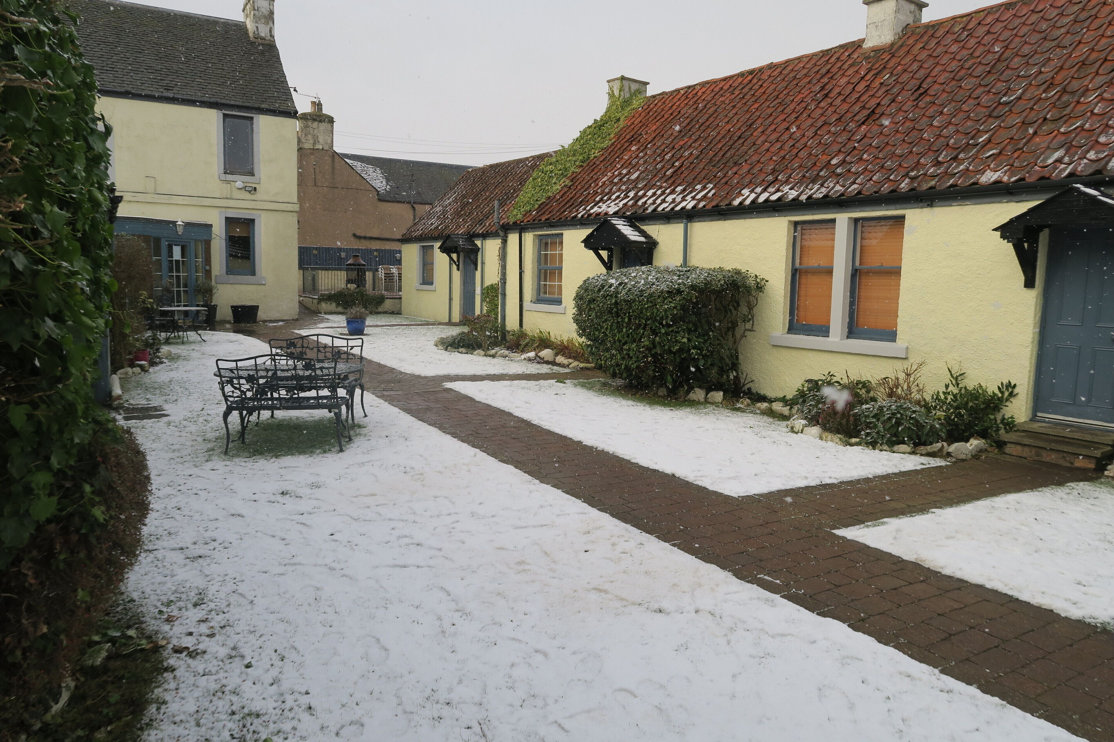 A snow-covered courtyard with a brick pathway, a bench, and a building with a red-tiled roof.
