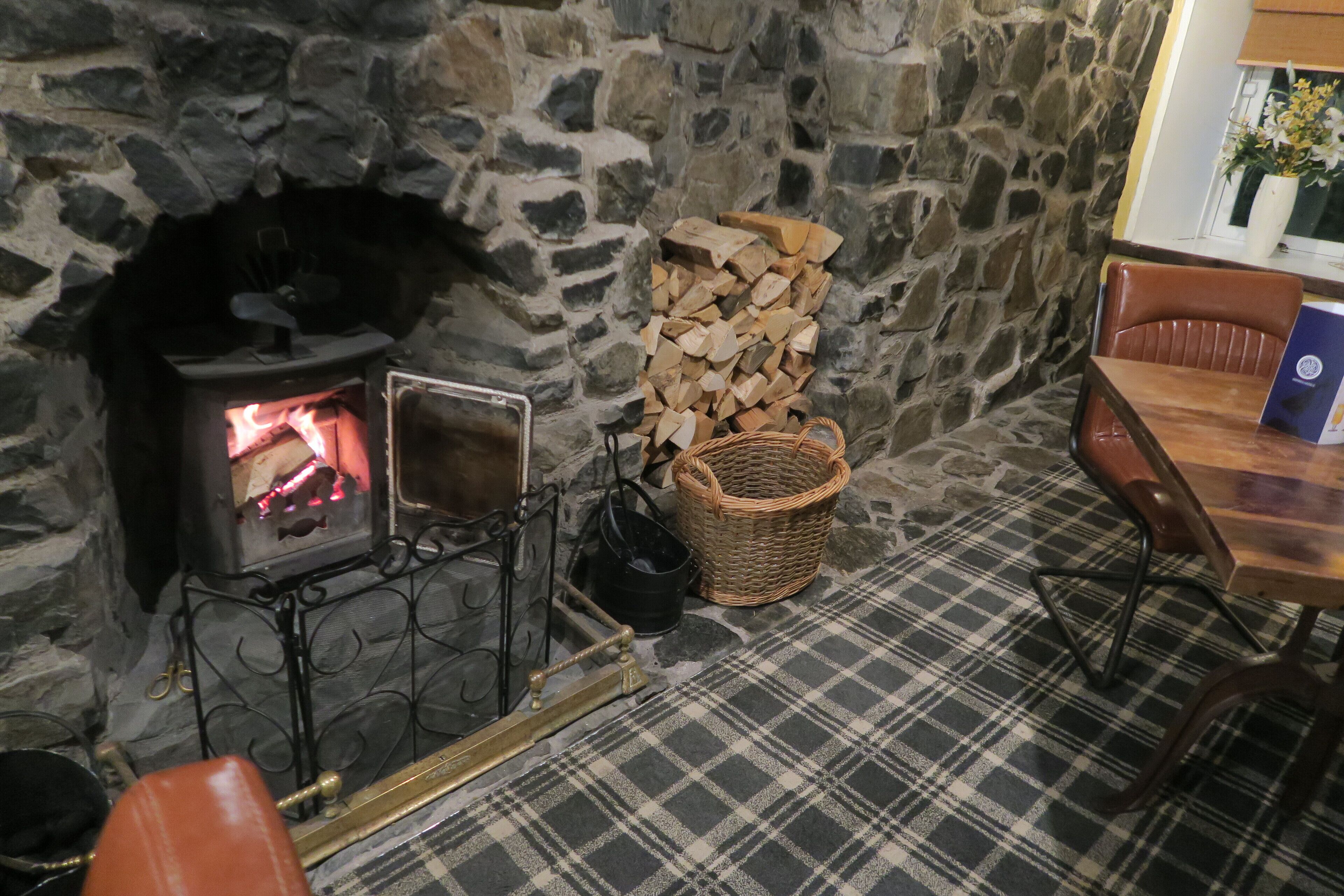 Une cheminée en pierre avec un poêle à bois, une pile de bois de chauffage, un panier en osier et un tapis à carreaux.