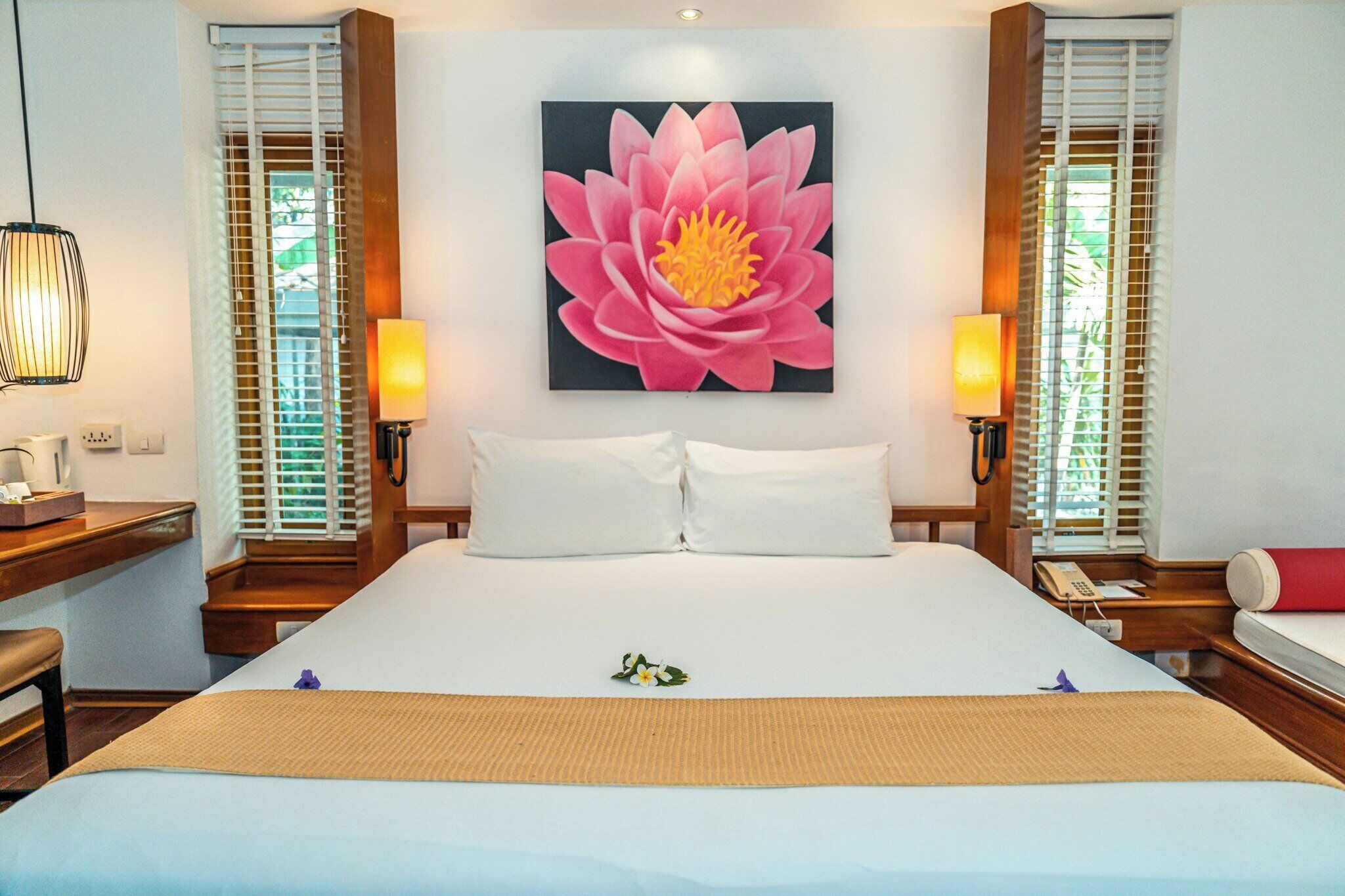 Quarto de hotel com uma cama grande, duas mesas de cabeceira, um criado-mudo, uma luminária na parede e uma grande obra de arte floral.
