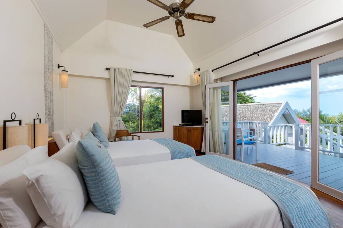 A bedroom with two beds, a ceiling fan, a TV, and a balcony with a view of the landscape.