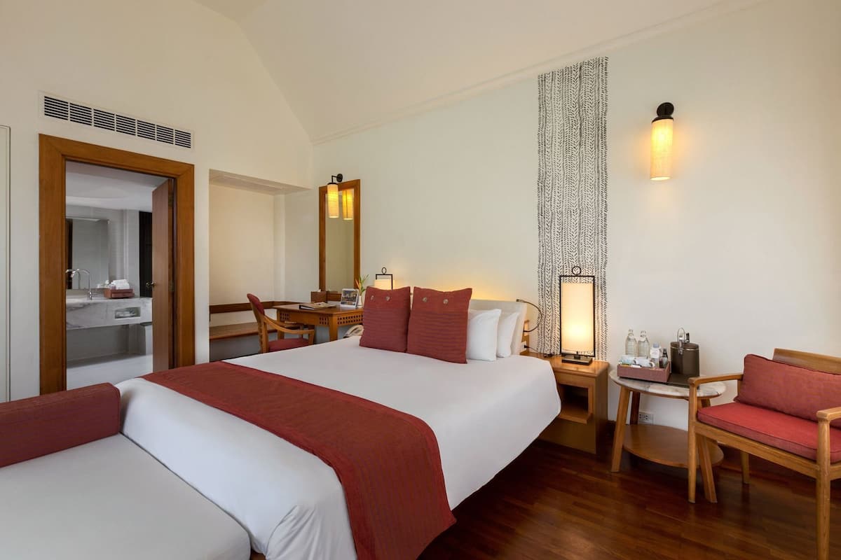 A hotel room with a large bed, a desk, a chair, a small table, a mirror, and a wall-mounted lamp.