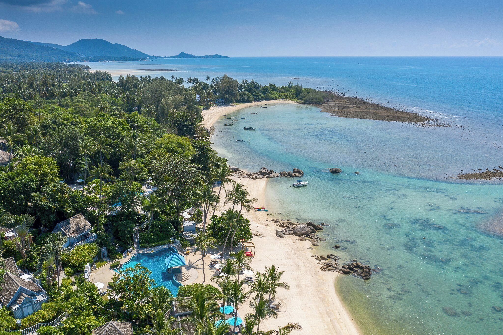 Vista aérea de um resort tropical com praia, águas cristalinas e vegetação exuberante.