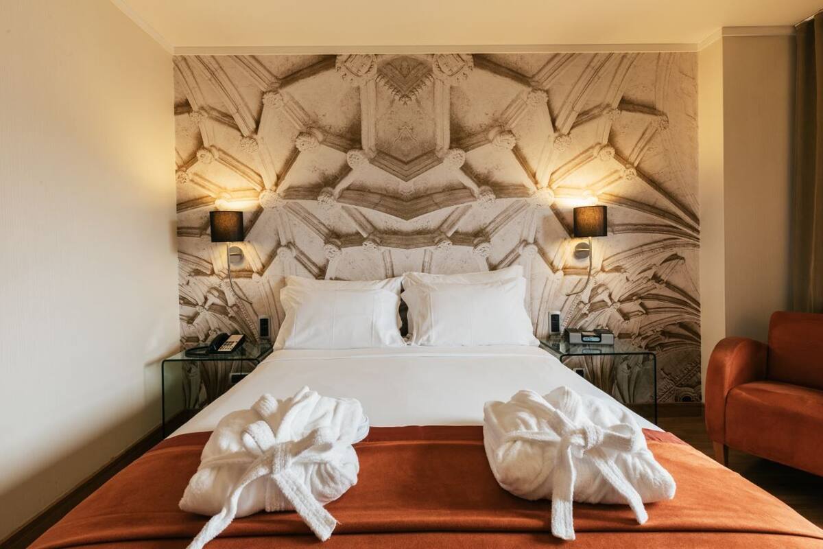 Une chambre d’hôtel avec un grand lit, deux tables de chevet, un canapé rouge et un mur décoratif orné d’un motif géométrique.
