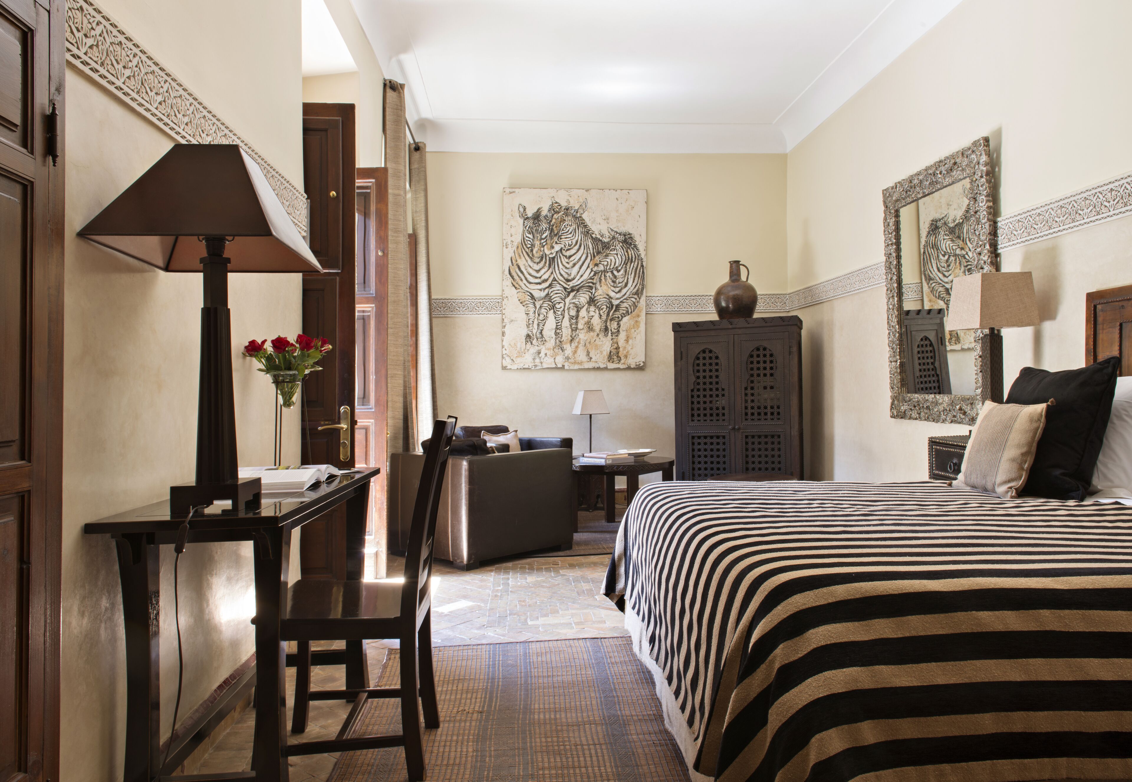 Habitación de hotel con cama, escritorio, silla, lámpara, jarrón con flores y una obra de arte en la pared.