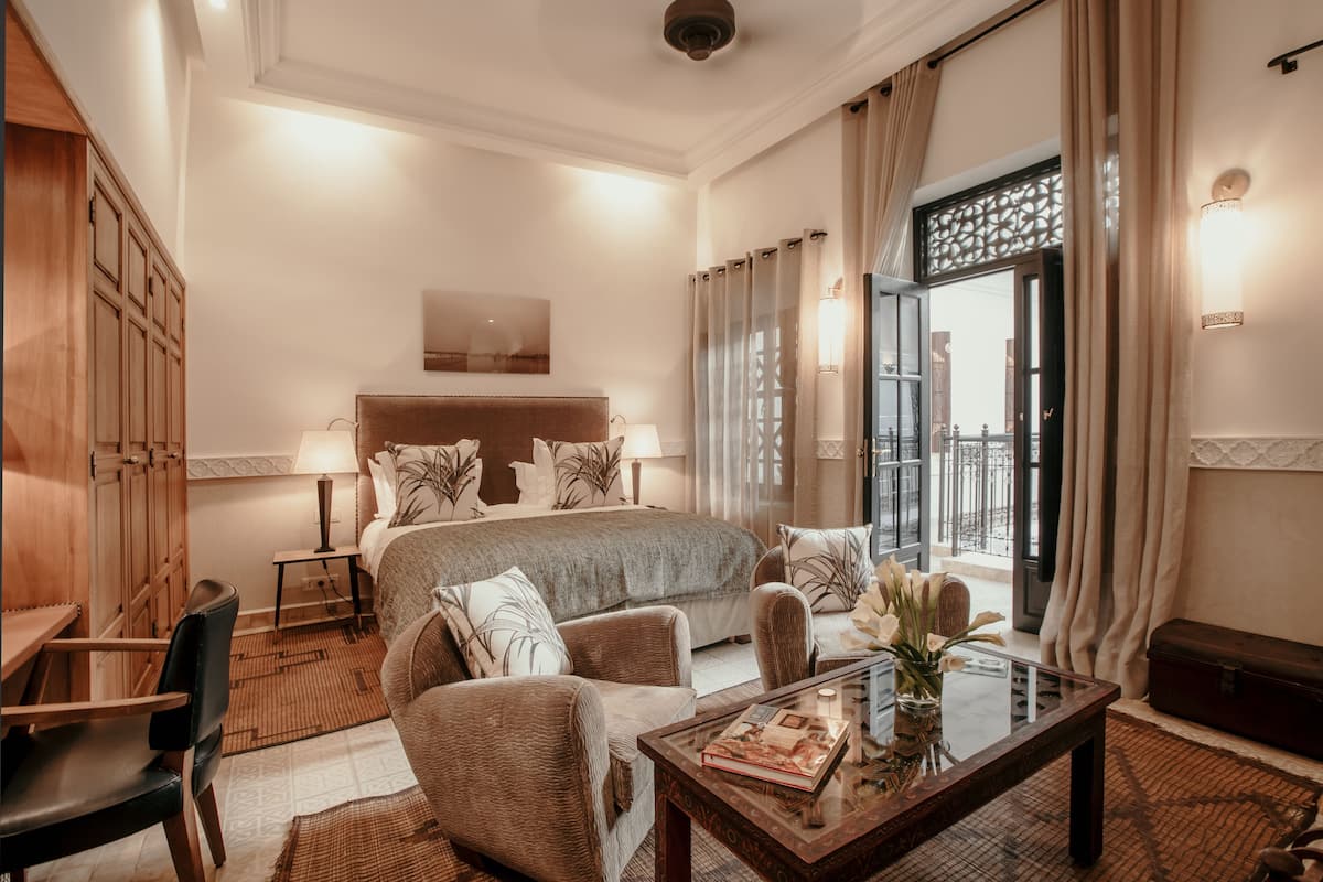A hotel room with a large bed, a sitting area with a sofa and armchairs, a small table with a book and flowers, and a view through a window with curtains.