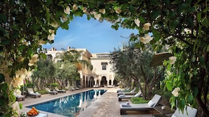 A poolside area with lounge chairs, a table with fruit, and a building with arches in the background. - La Villa Des Orangers - Relais & Châteaux (Marrakech)