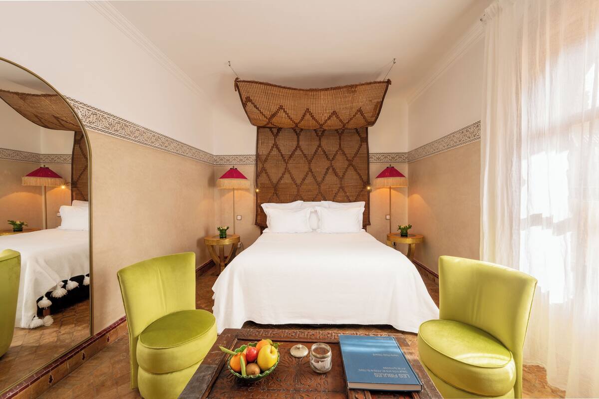 A hotel room with a large bed, two green chairs, a small table with fruit and a book, and two lamps.